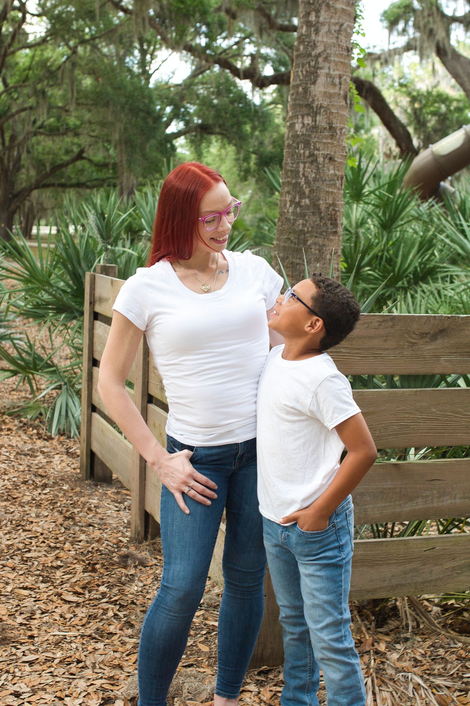 Red-haired woman and young boy sharing a joyful moment by a wooden fence in a lush park.