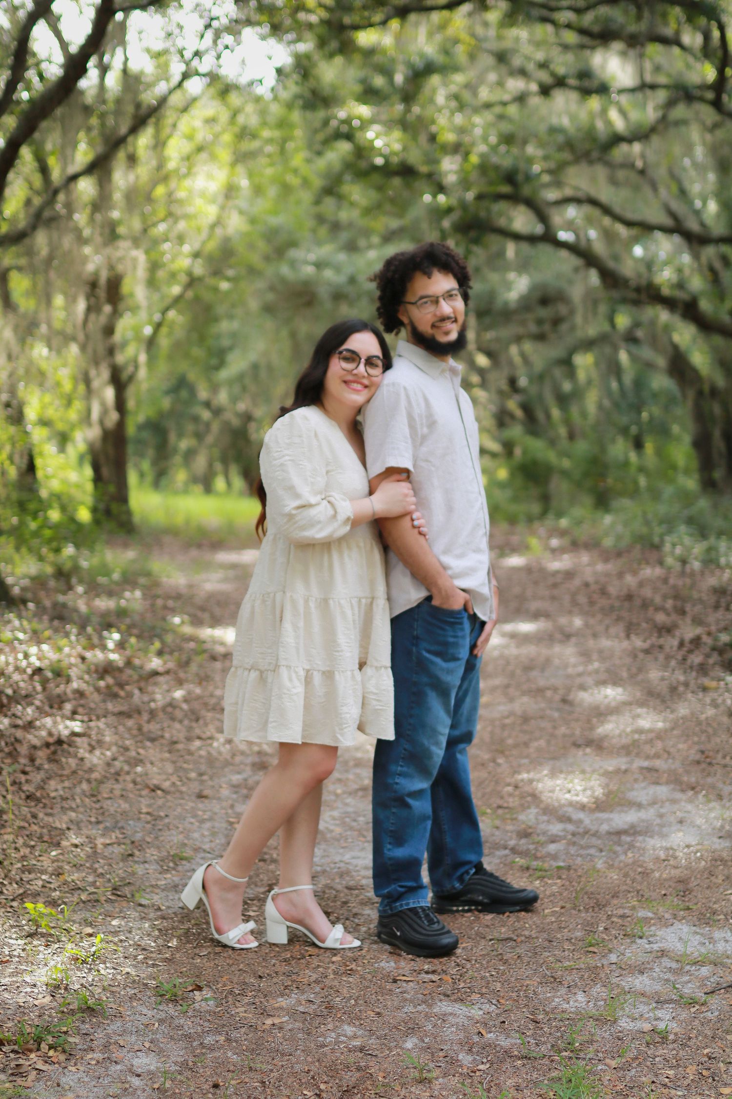 A couple stands together on a wooded path, wearing white and denim while embracing in natural lighting.
