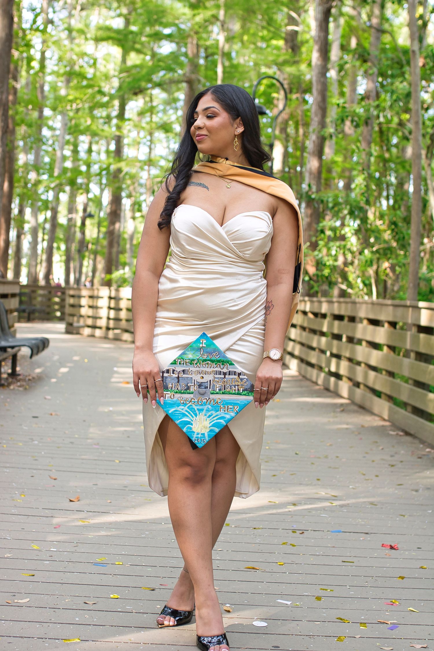 Woman in cream satin dress holding decorated graduation cap on wooden boardwalk surrounded by lush green trees.