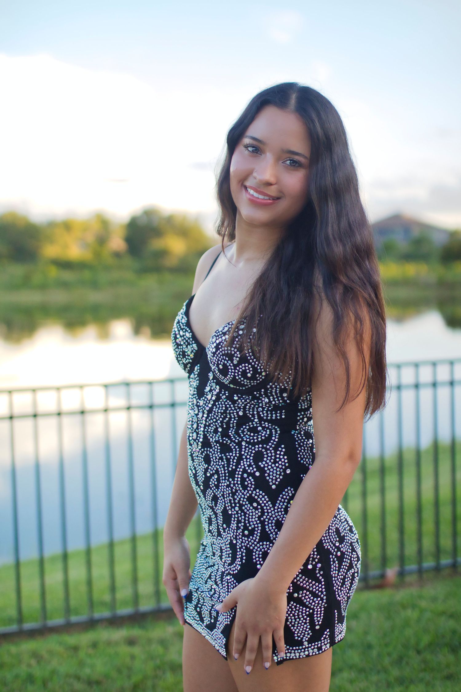 Young woman in black beaded mini dress smiling outdoors near a lake at sunset.