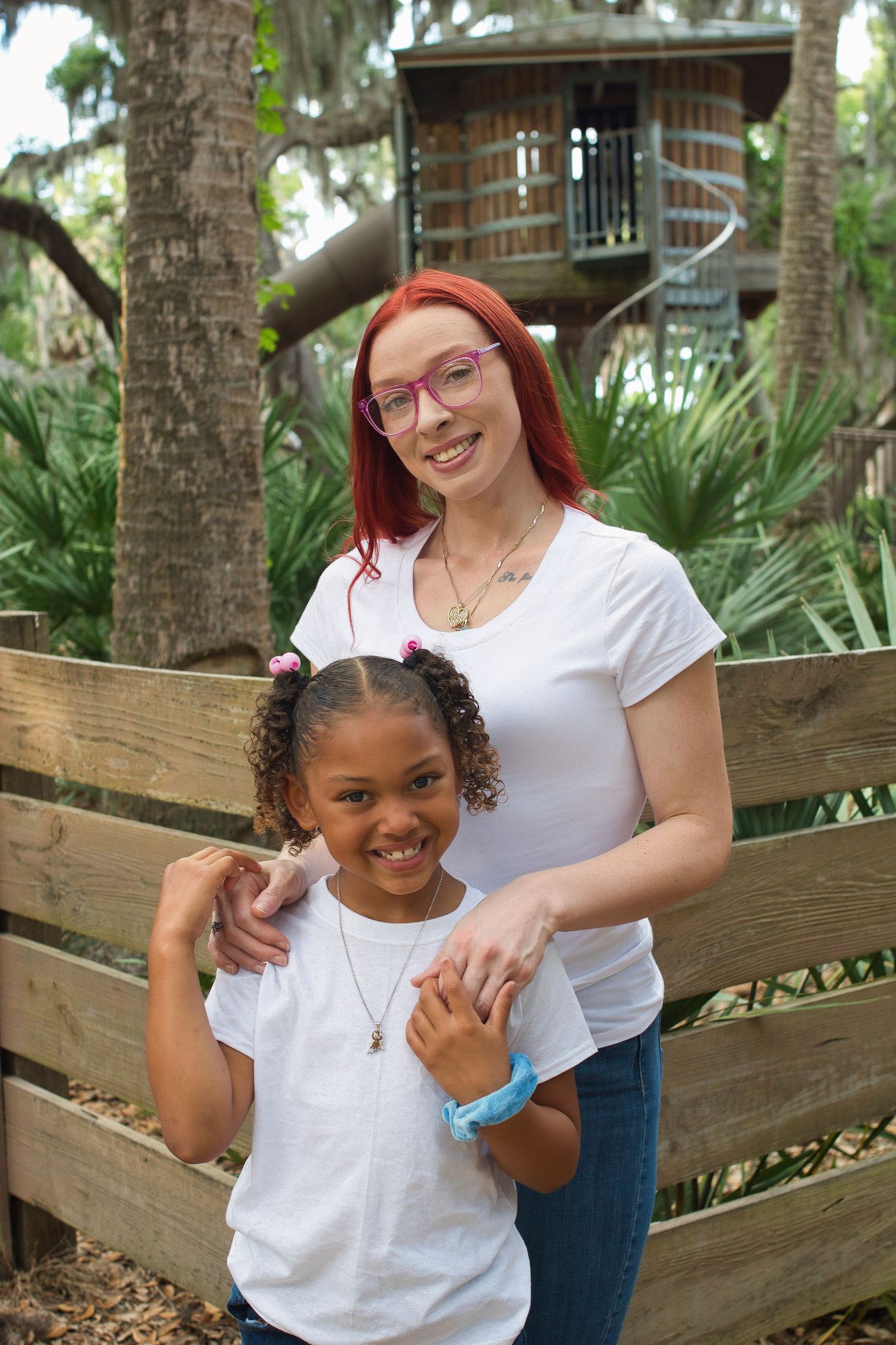 Woman with red hair and young girl in white shirts smiling near wooden fence outdoors.