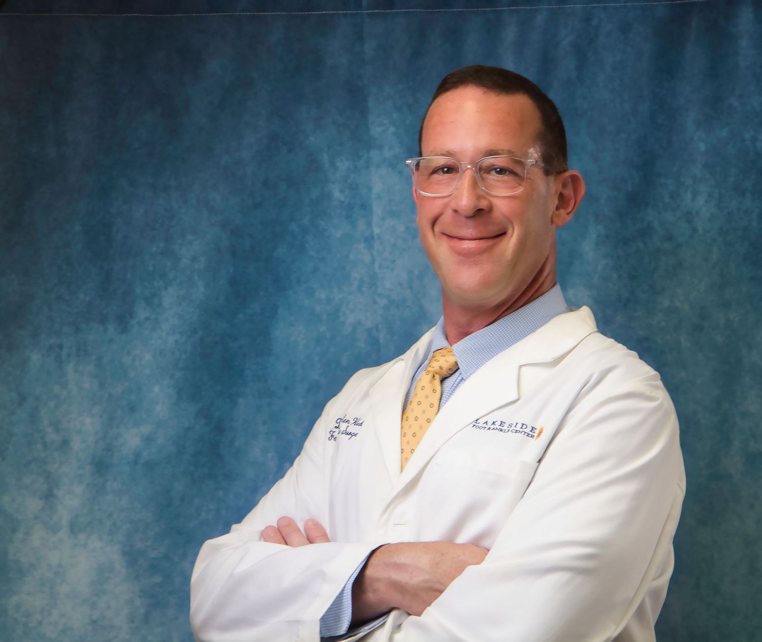 Smiling male doctor in white lab coat with arms crossed against blue background.