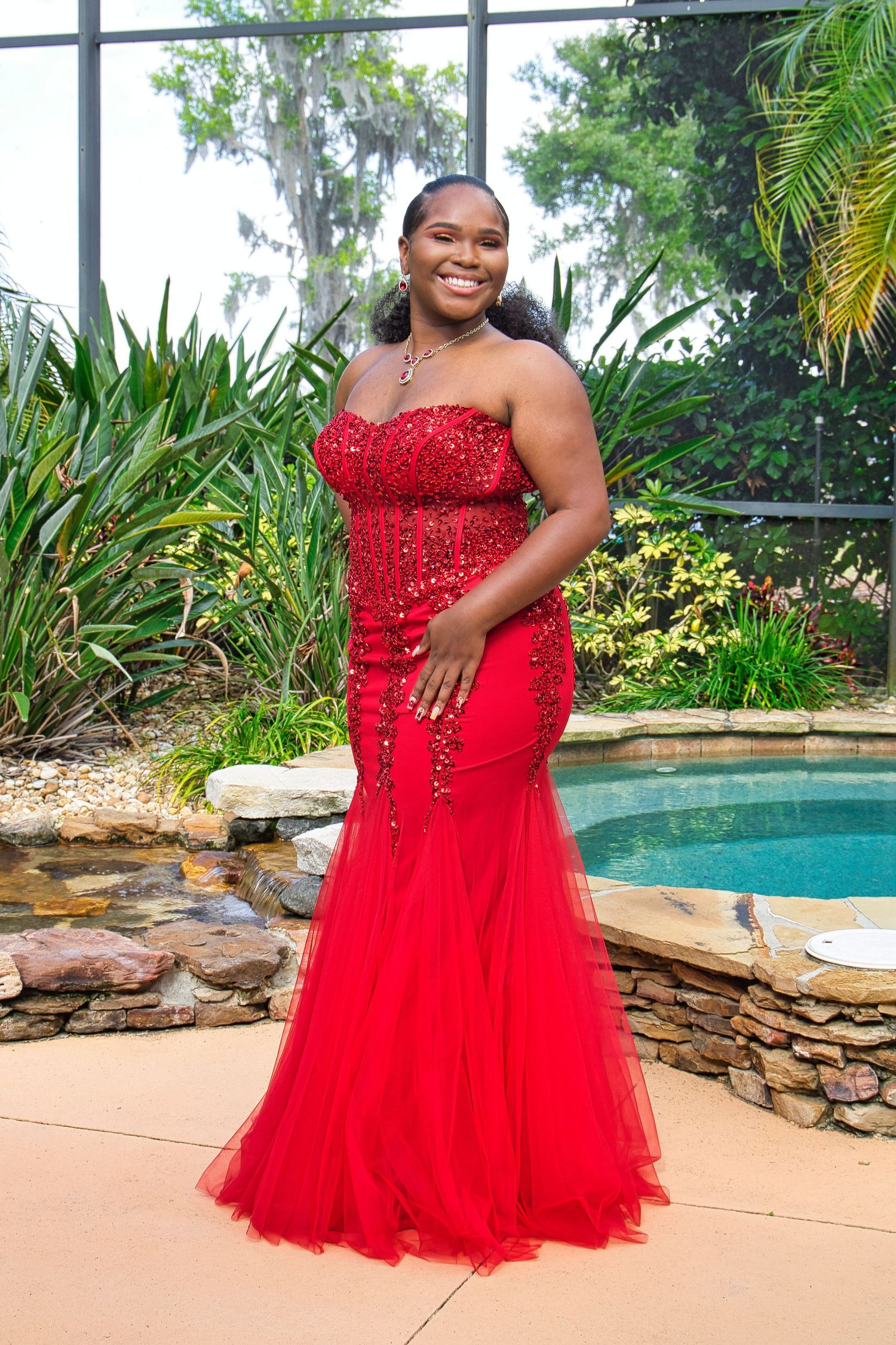 Elegant strapless red gown with sequins and tulle photographed in an outdoor garden setting.