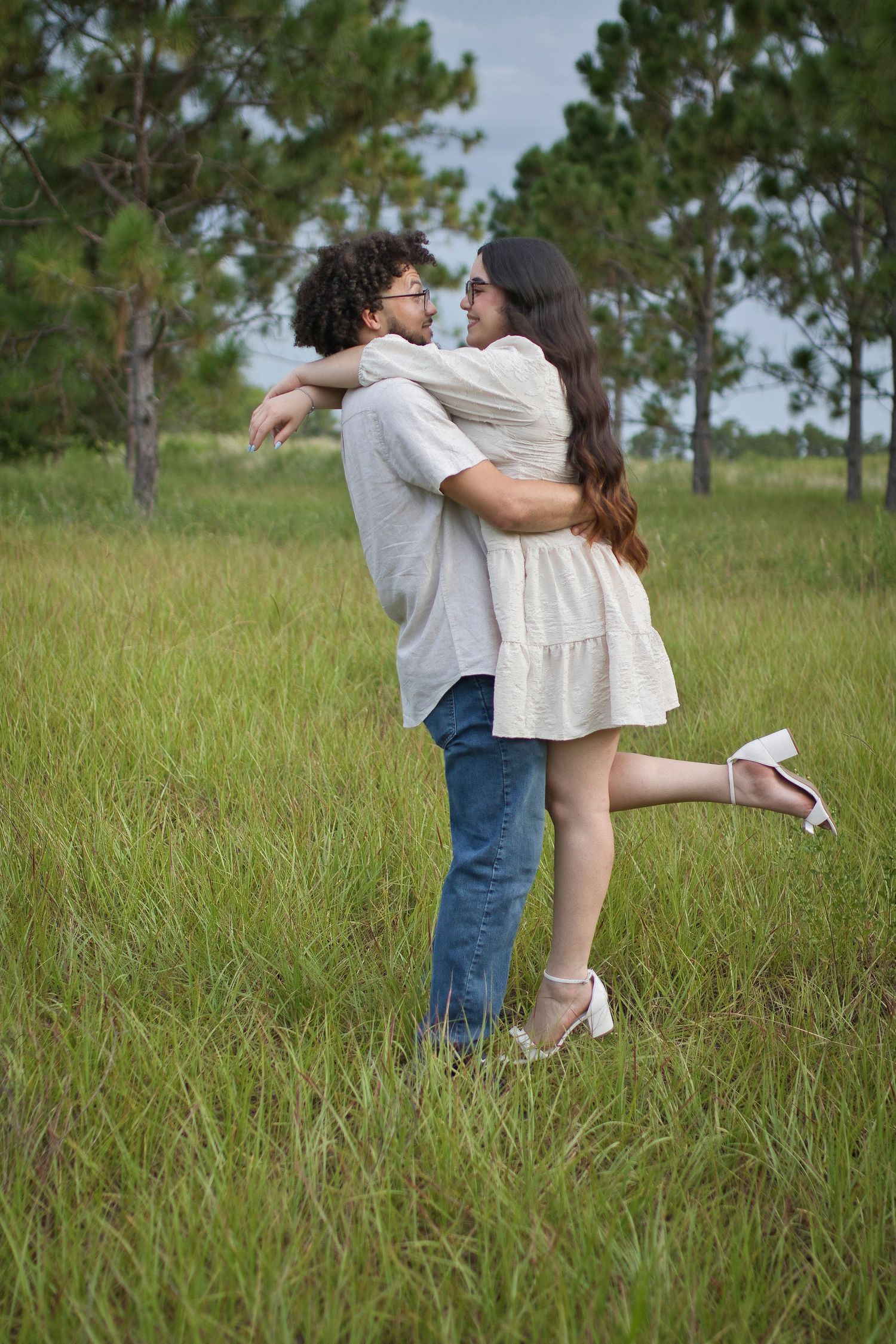 A romantic couple embraces in a sunlit meadow with tall grass and pine trees in the background.