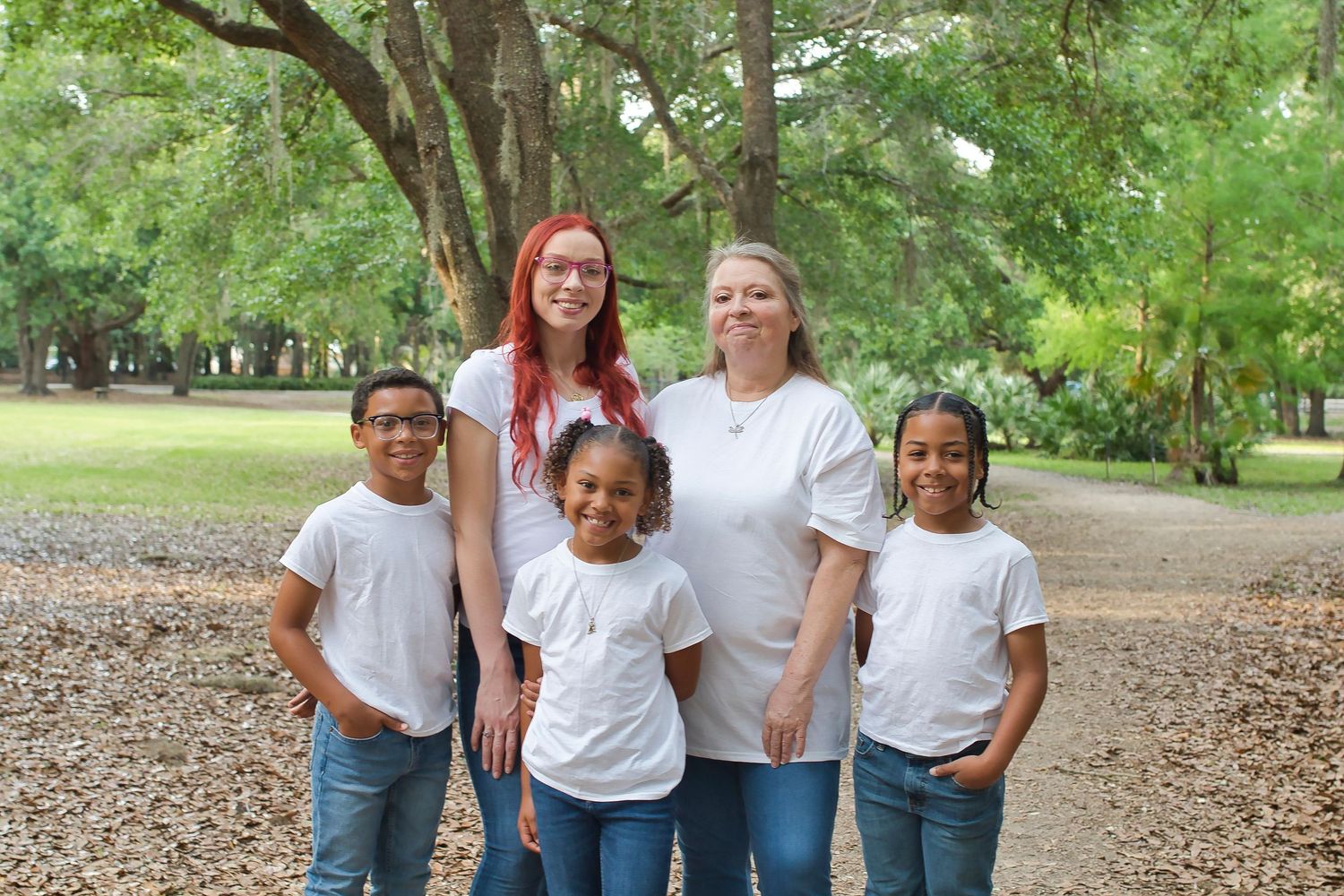 Family of five posing outdoors in matching white shirts and jeans among trees in a park.