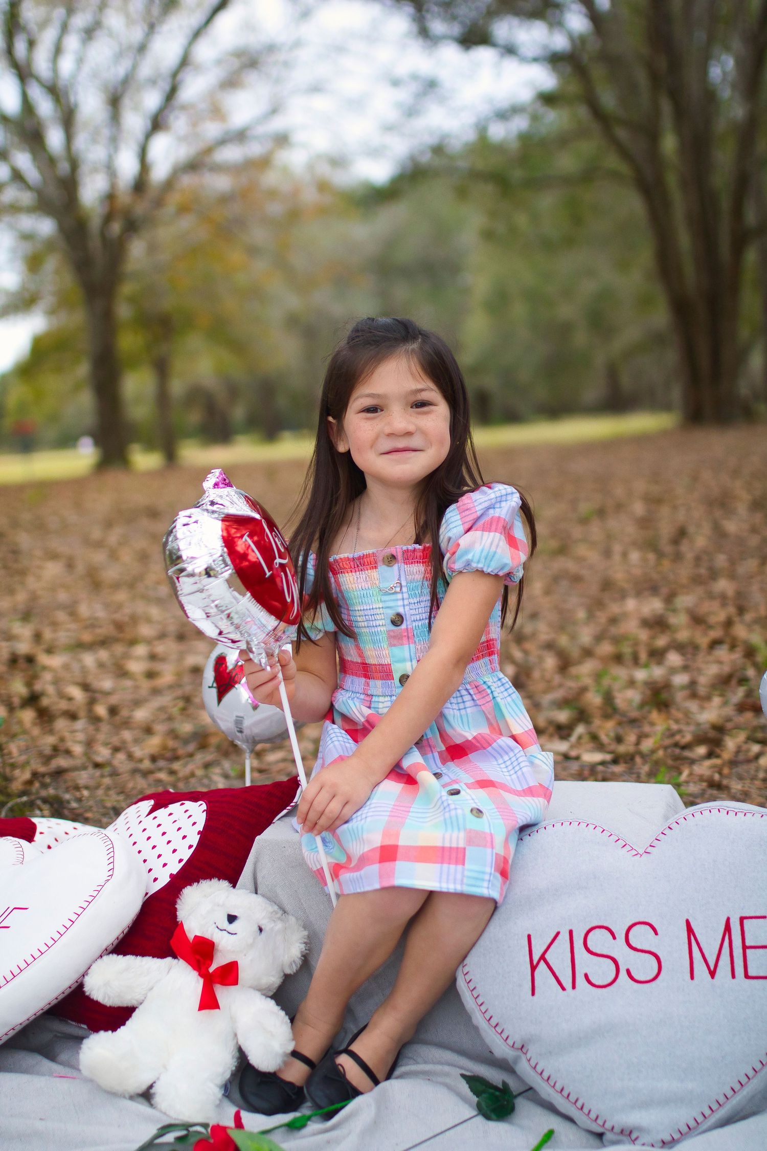 Young child in plaid dress poses with Valentine's heart pillows and stuffed bears outdoors.