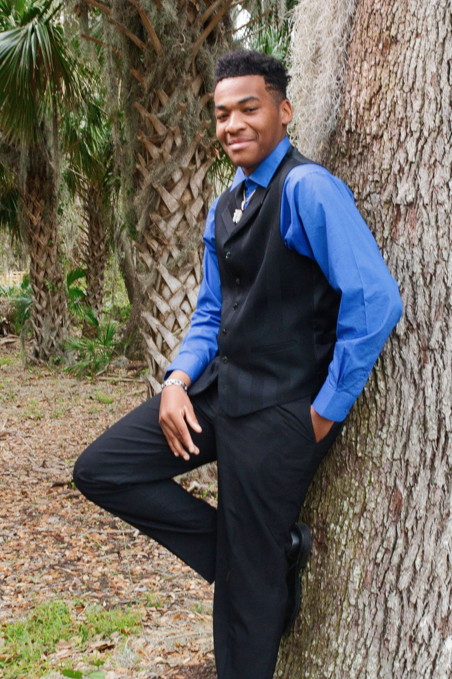 Young man in blue shirt and black vest smiling while leaning against a tree outdoors.