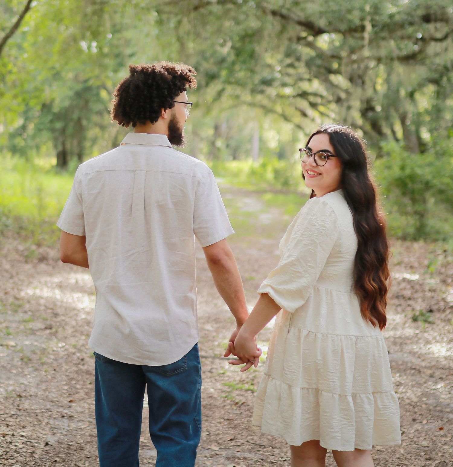 Couple holding hands on a forest path, woman in white dress smiling back at camera.