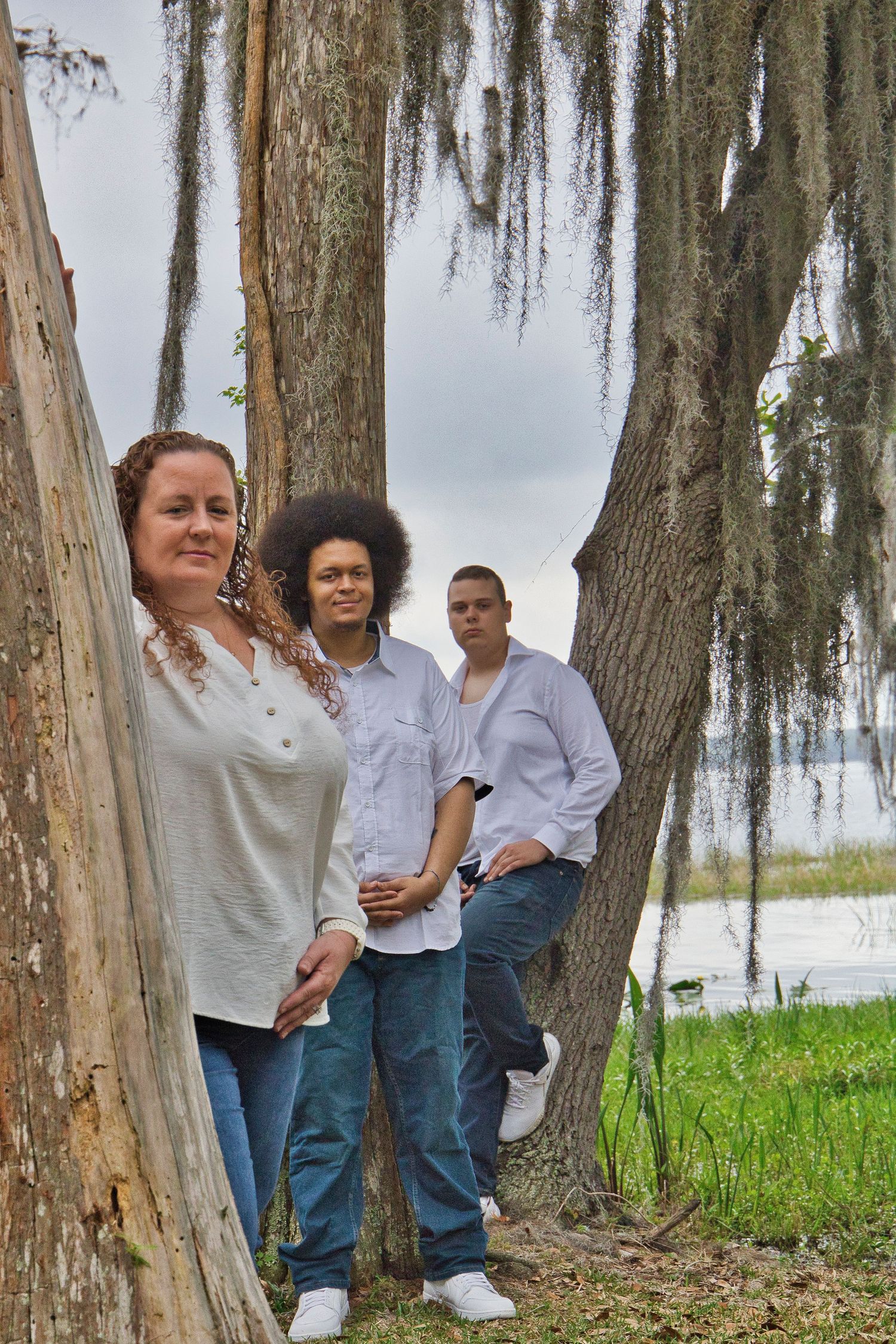 Three people posing among moss-draped trees near a lake, wearing casual jeans and button-up shirts outdoors.