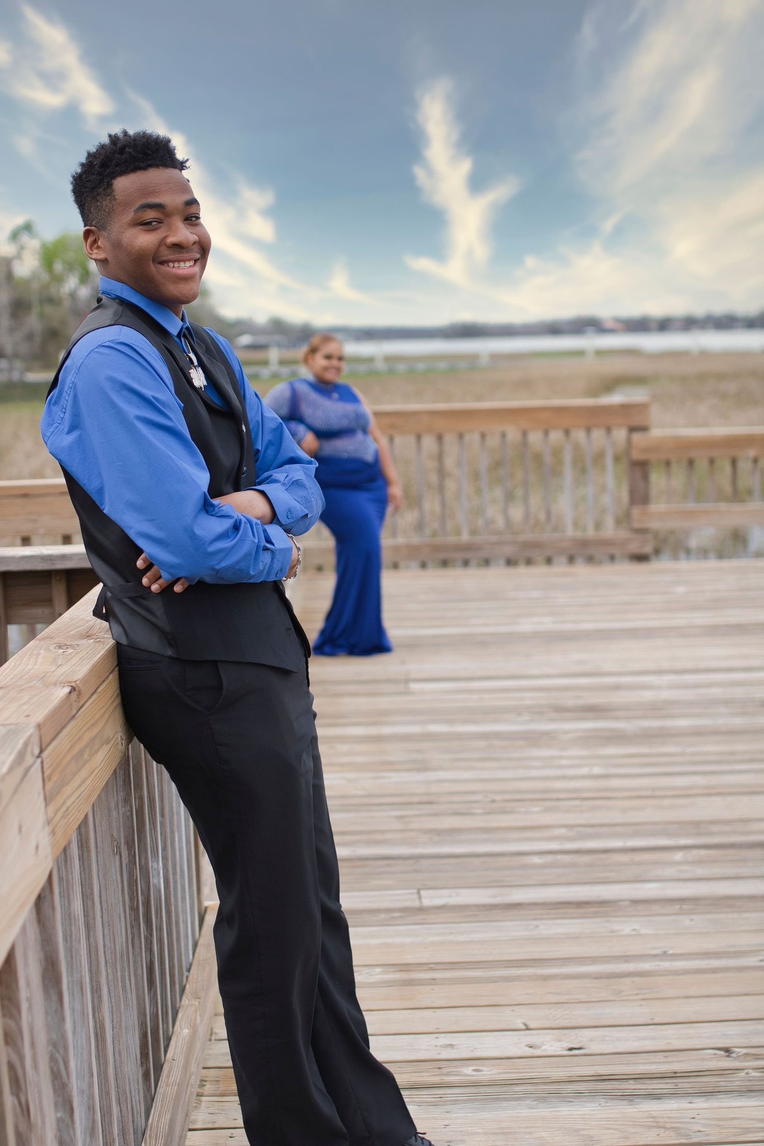 A smiling young man in a blue shirt and black vest poses on a wooden boardwalk with a woman in a blue gown behind him.