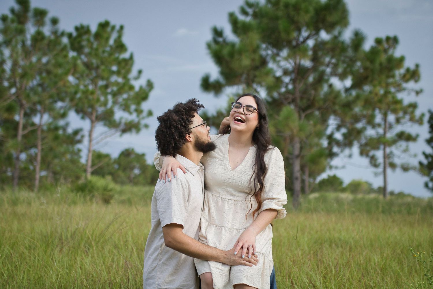Happy couple laughing and embracing in a green meadow with tall pine trees in the background.