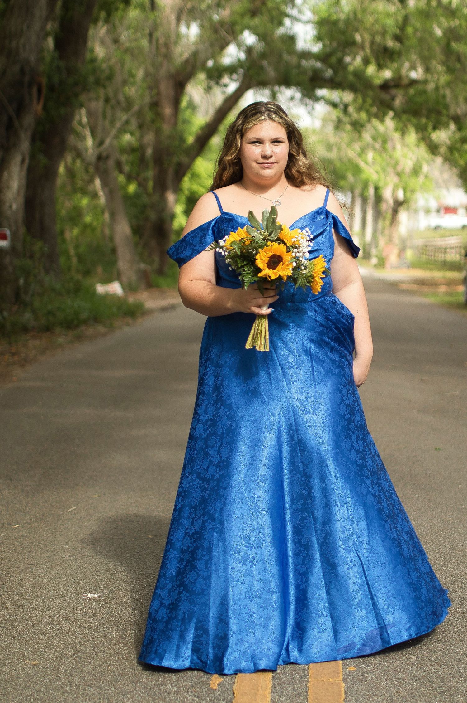 Woman in blue off-shoulder gown holding sunflower bouquet standing on tree-lined road.