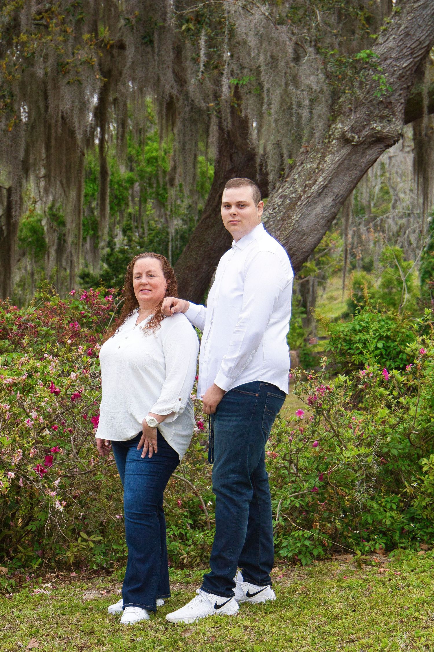 A woman and tall young man in white shirts and jeans pose outdoors near a moss-draped oak tree with blooming azaleas.