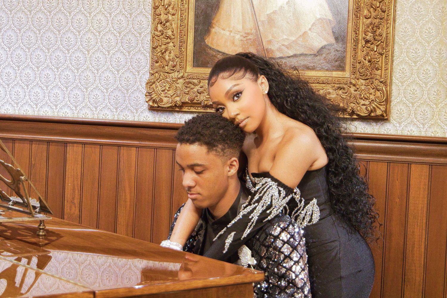 A couple poses at a grand piano, woman in sequined top leans over seated man in elegant wood-paneled room.