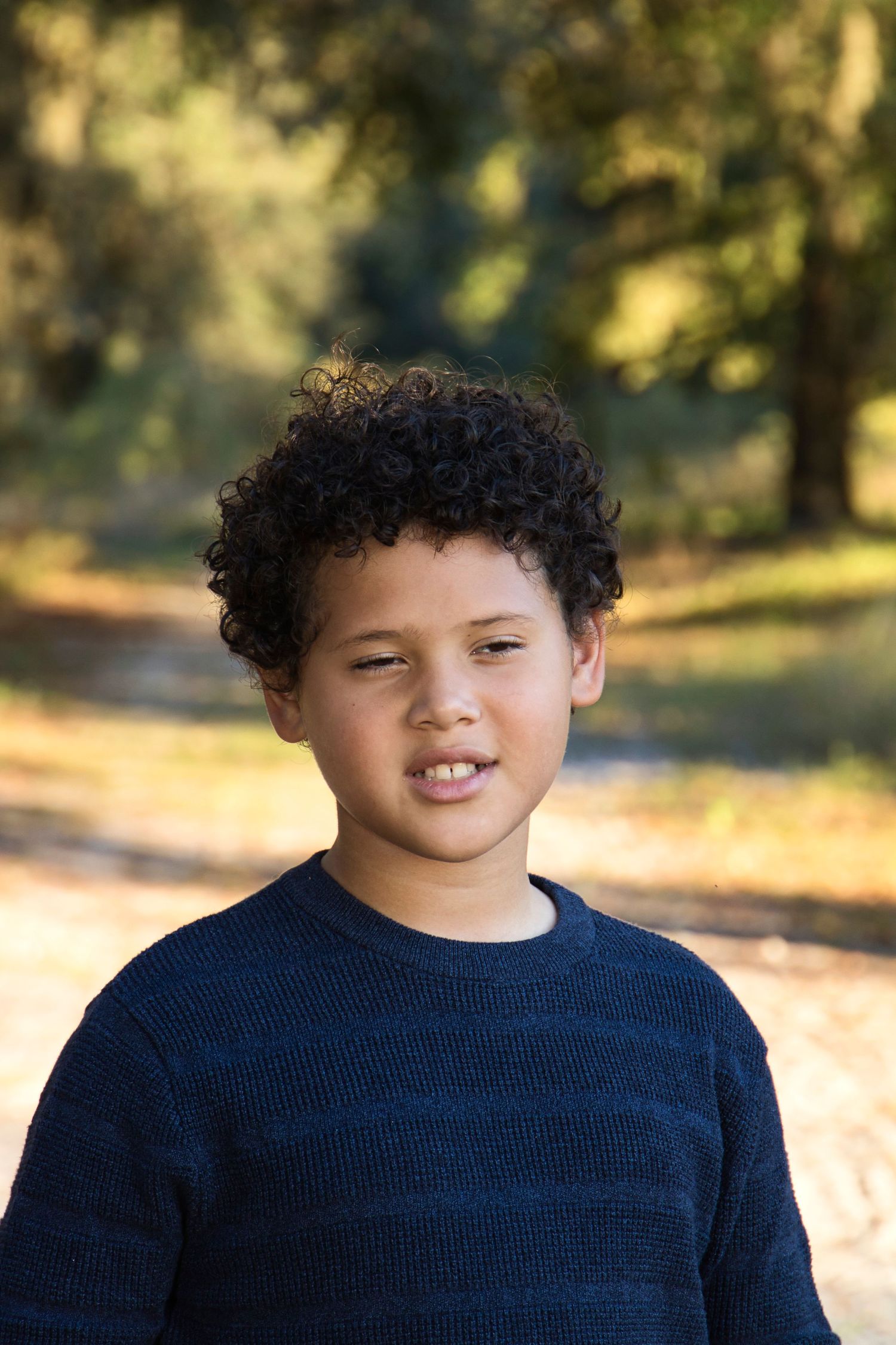 A young boy with curly hair wearing a navy sweater smiles outdoors in a sunlit park setting.