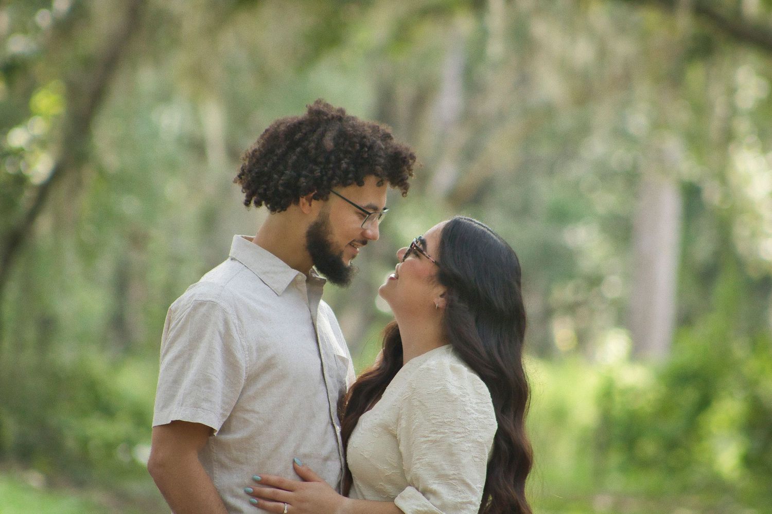 A couple shares a romantic moment outdoors, gazing at each other in a lush green forest setting.