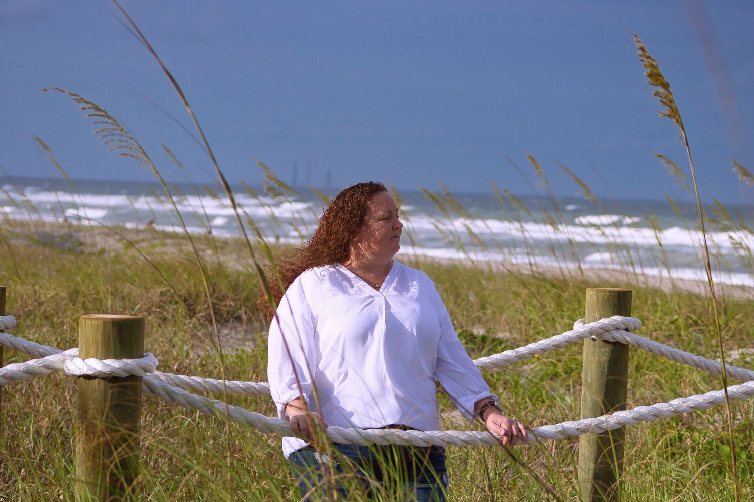 Person in white shirt standing by beach fence with ocean waves and sea grass in background.