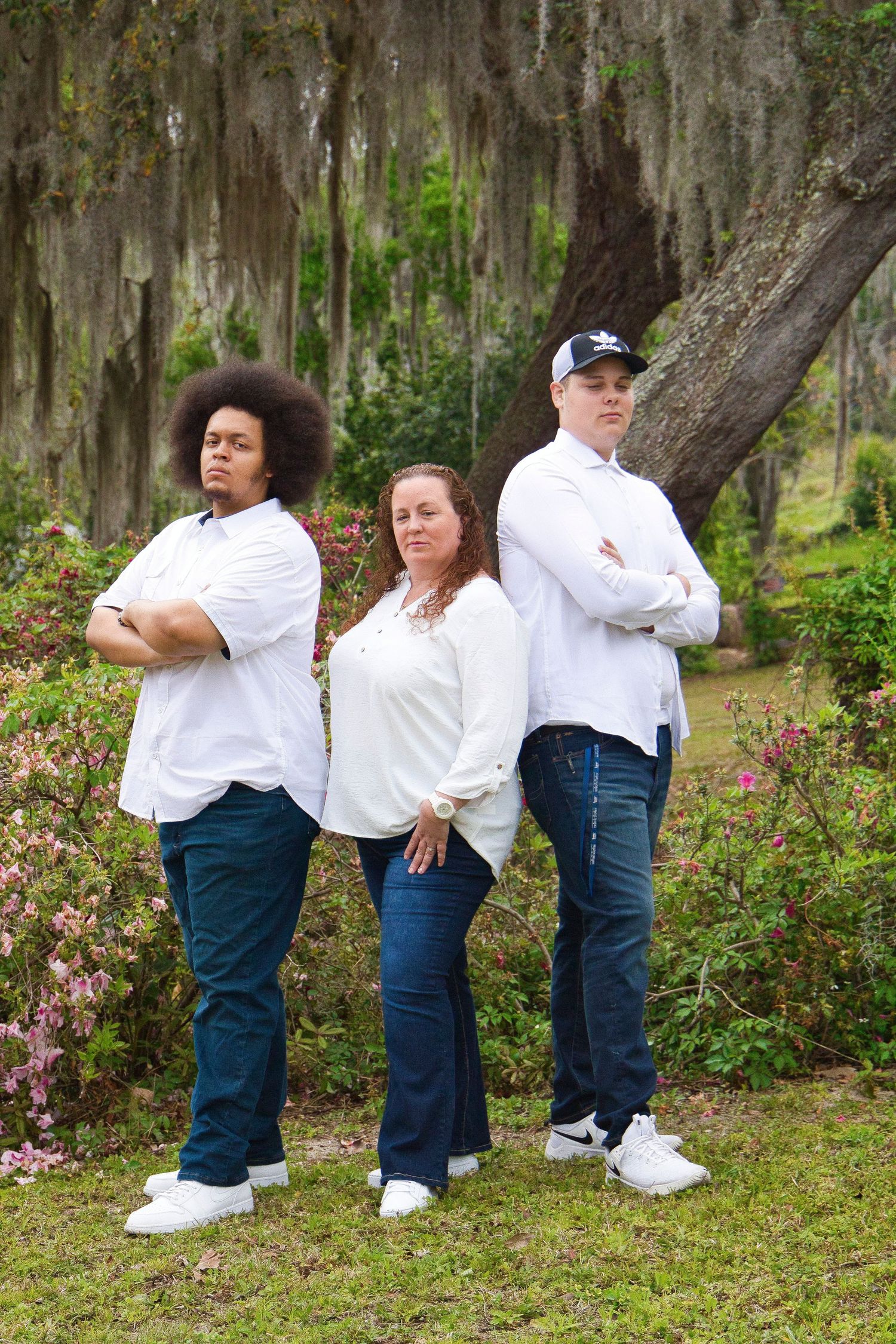 Three people in white shirts and jeans pose with arms crossed outdoors near a moss-covered tree with pink flowers.