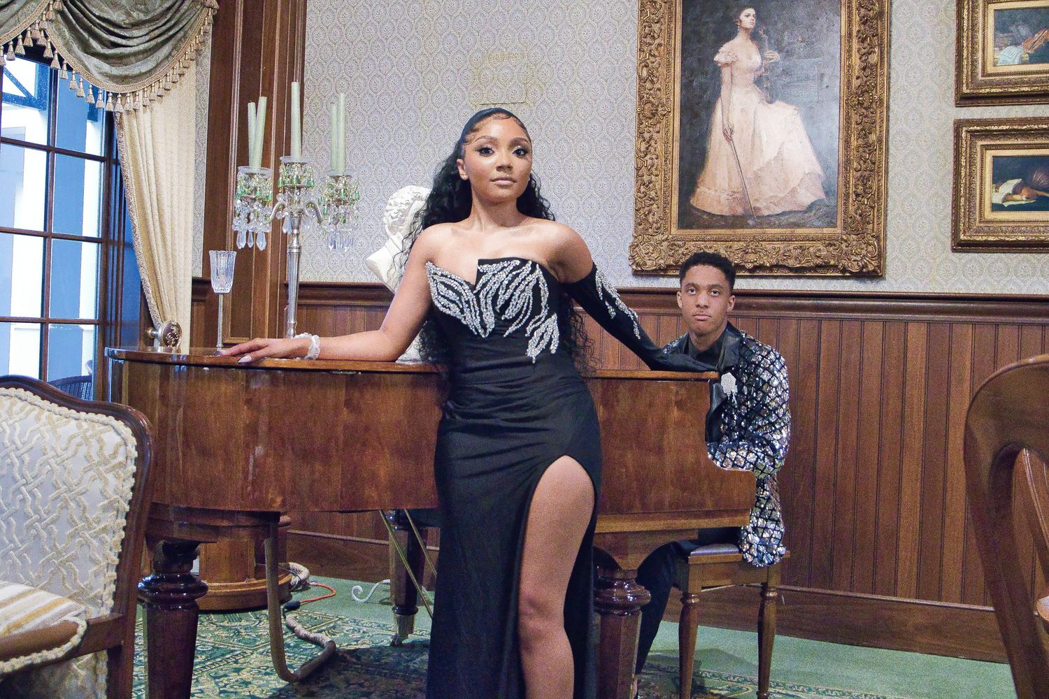 Woman in black zebra-print gown poses by piano while man in floral suit sits behind her in elegant room.
