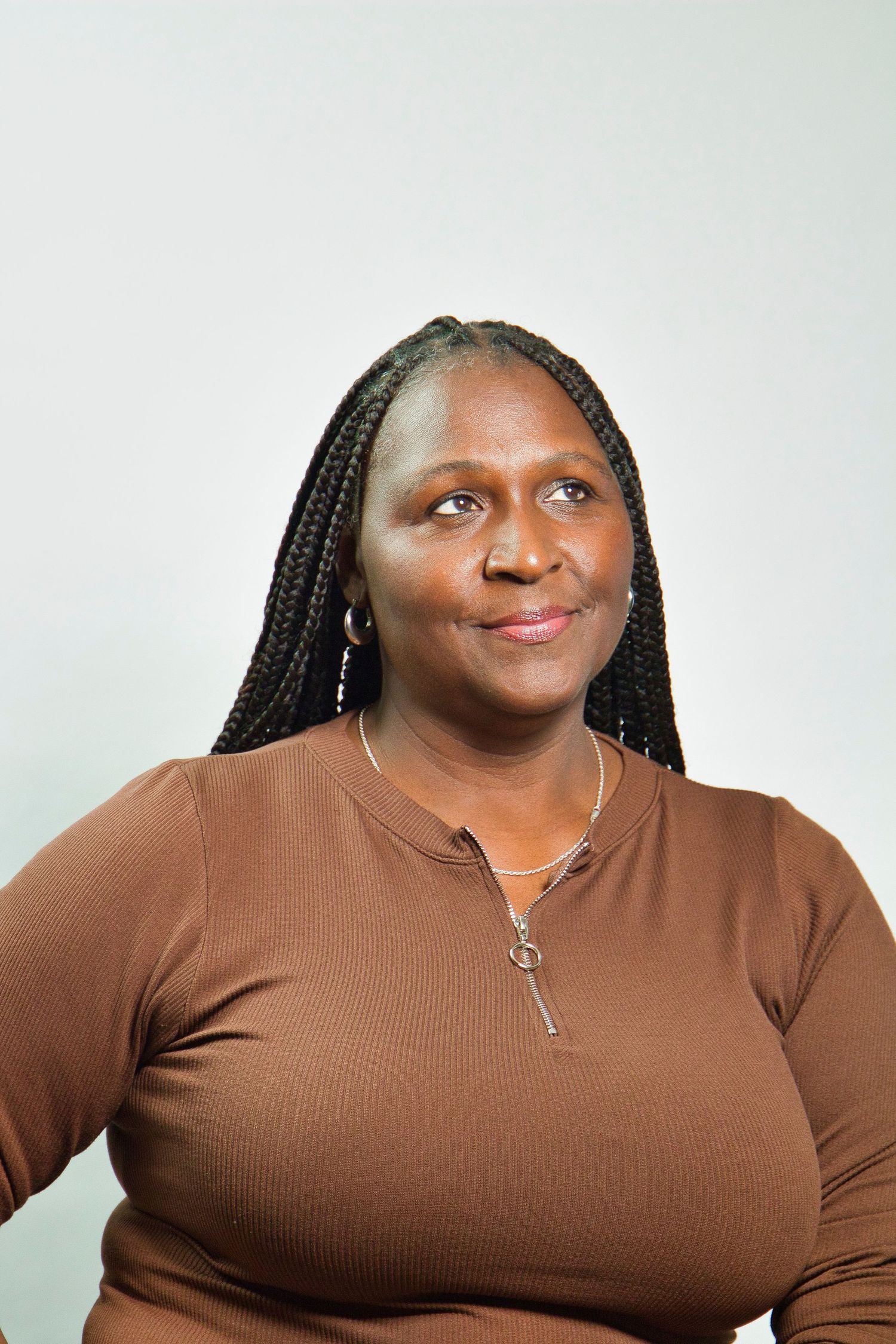 A woman with long braids wearing a brown top and necklace smiles confidently against a light gray background.