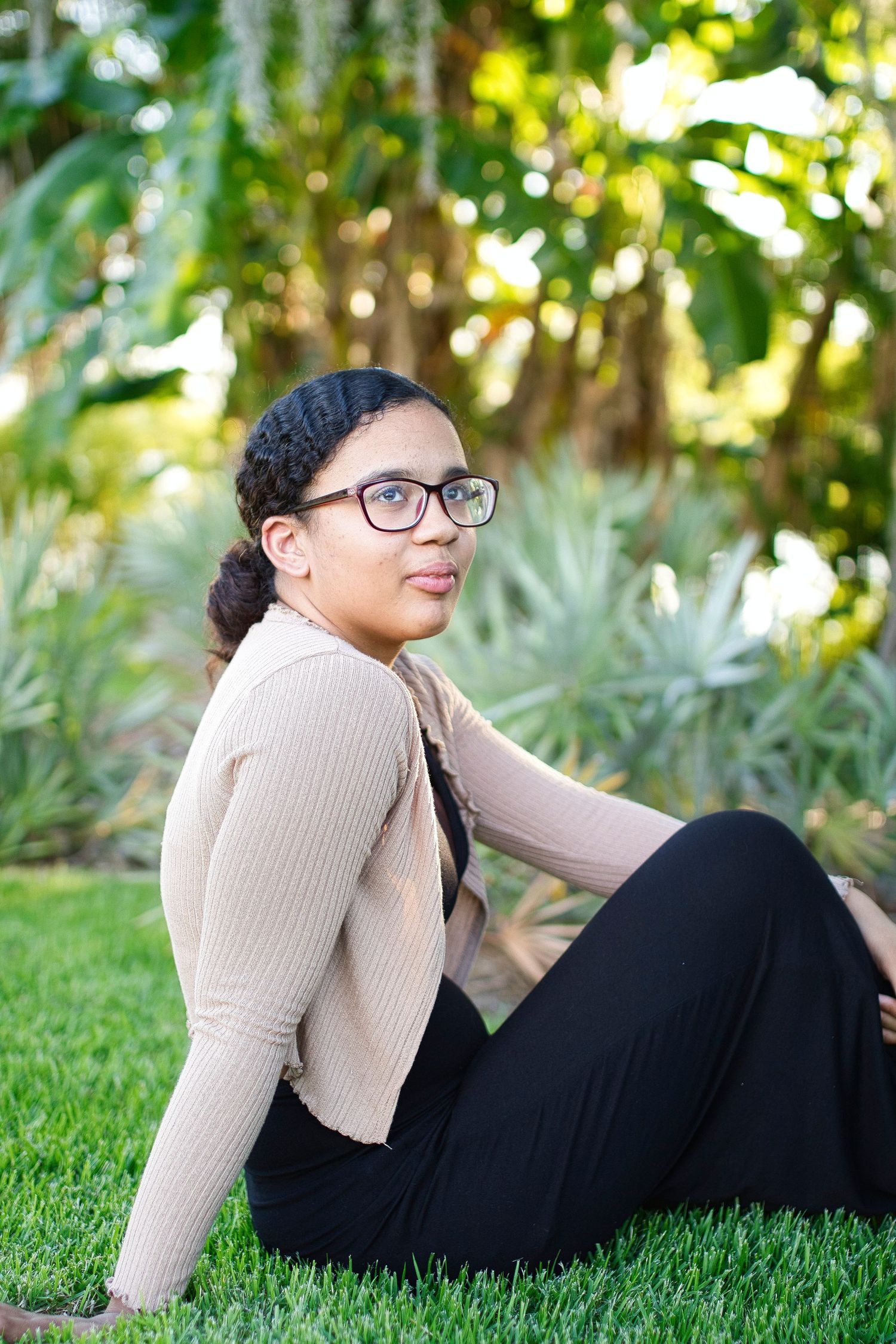 Young woman with braids and glasses in beige cardigan sitting on grass in a tropical garden setting.