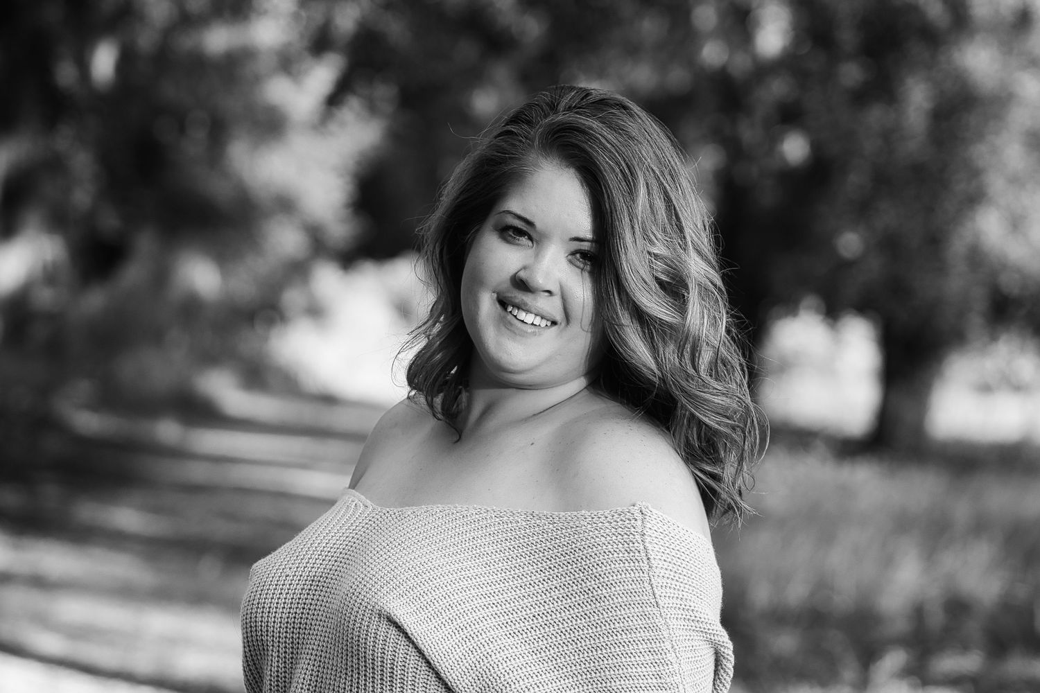 Black and white portrait of a smiling young woman with wavy hair wearing an off-shoulder sweater outdoors.