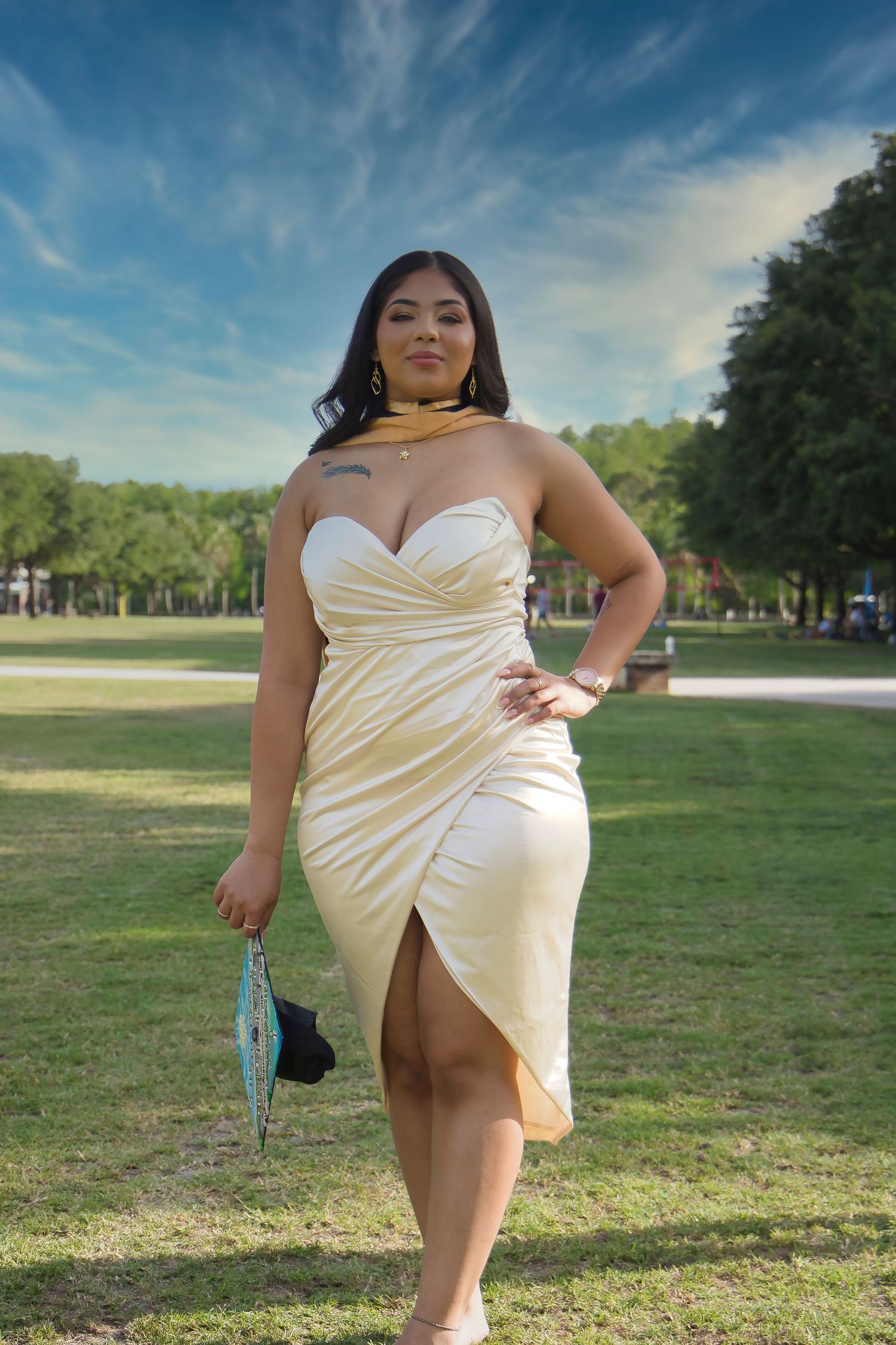 A woman in a cream-colored dress poses outdoors in a grassy park area on a sunny day.