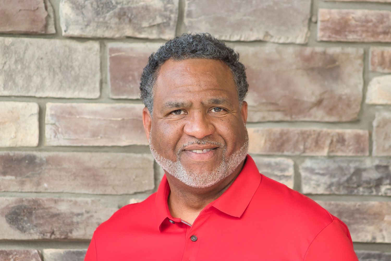 Smiling middle-aged man with gray beard wearing a red polo shirt, posed against a stone wall background.