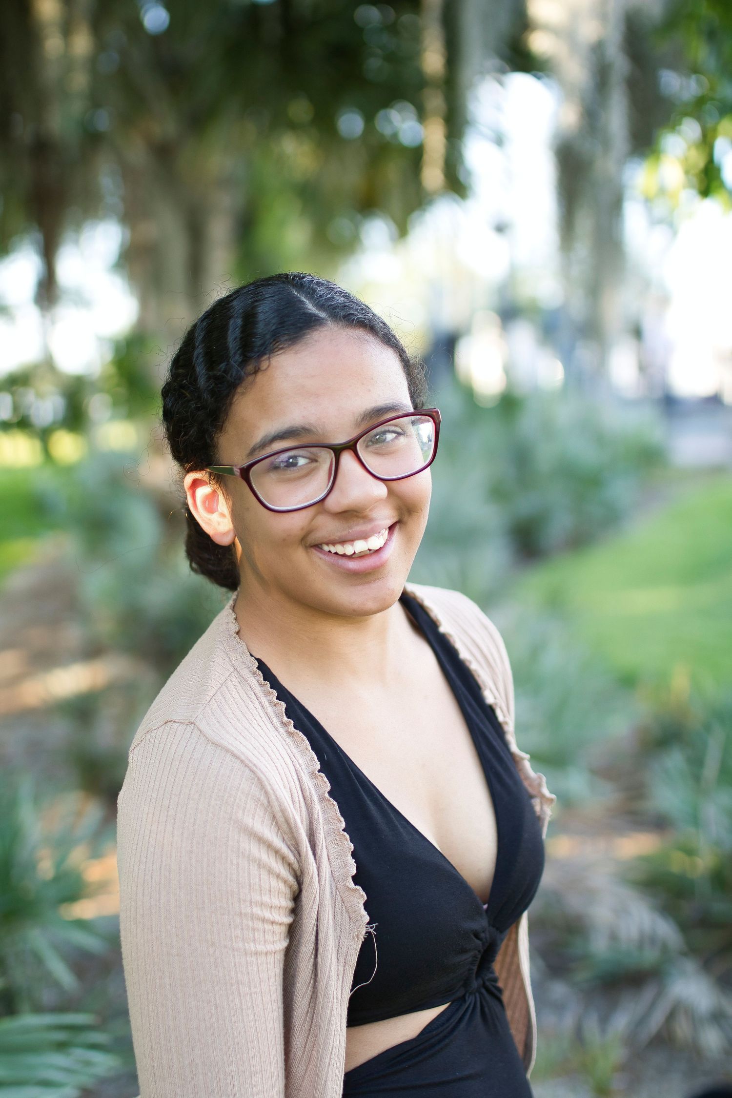 Smiling young woman with braids and glasses wearing a beige cardigan outdoors in a lush green park.