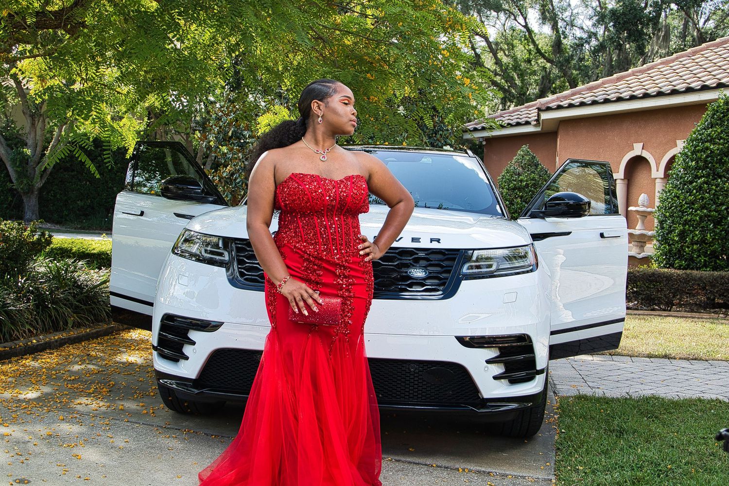 Red mermaid gown photographed in front of a white luxury vehicle and garden backdrop.