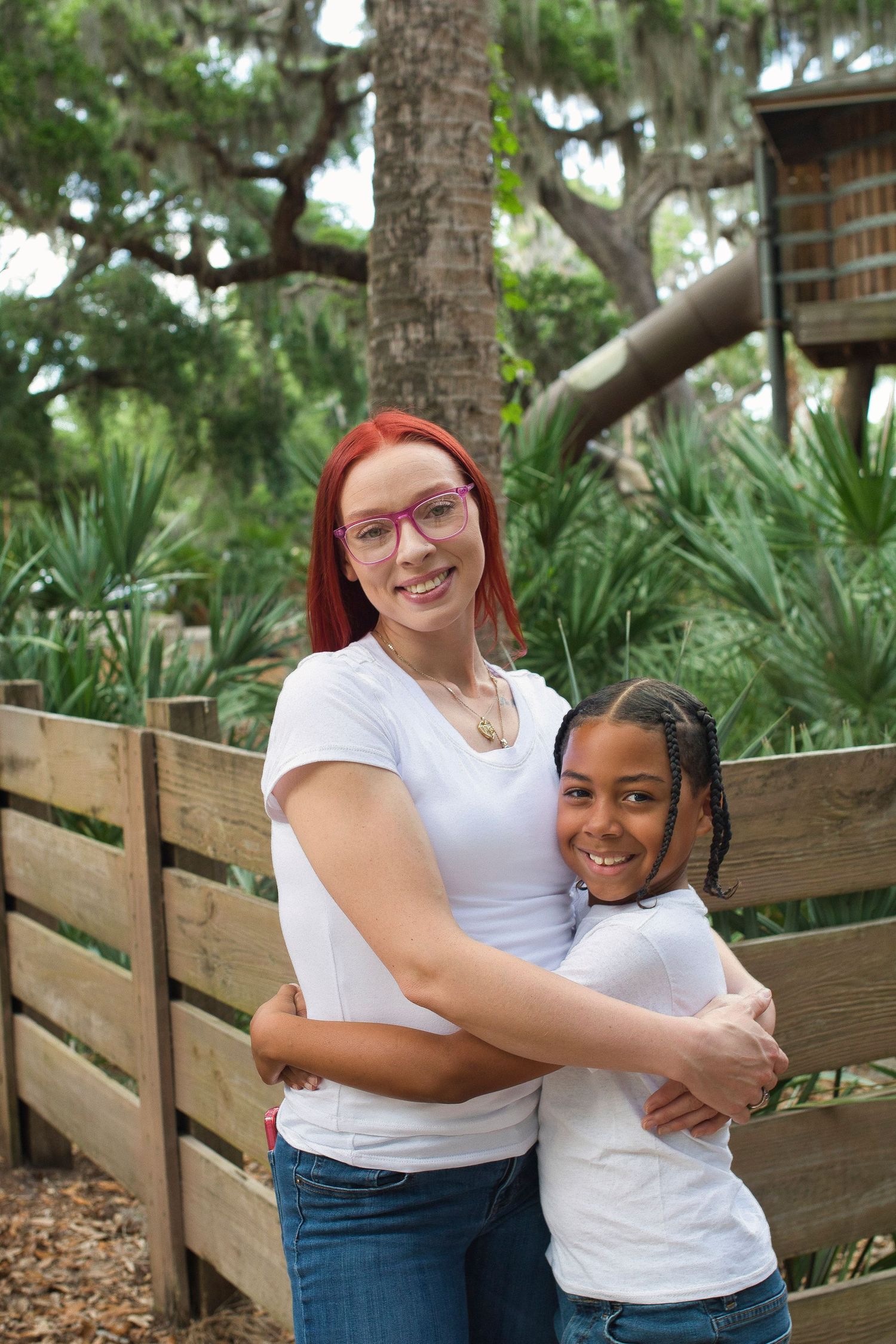 Woman with red hair hugging a young girl outdoors near a wooden fence in a green park.