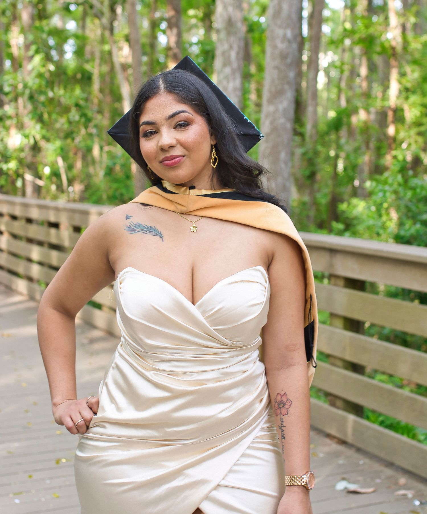 Woman in white satin dress and graduation cap and gold stole poses on wooden bridge surrounded by trees.