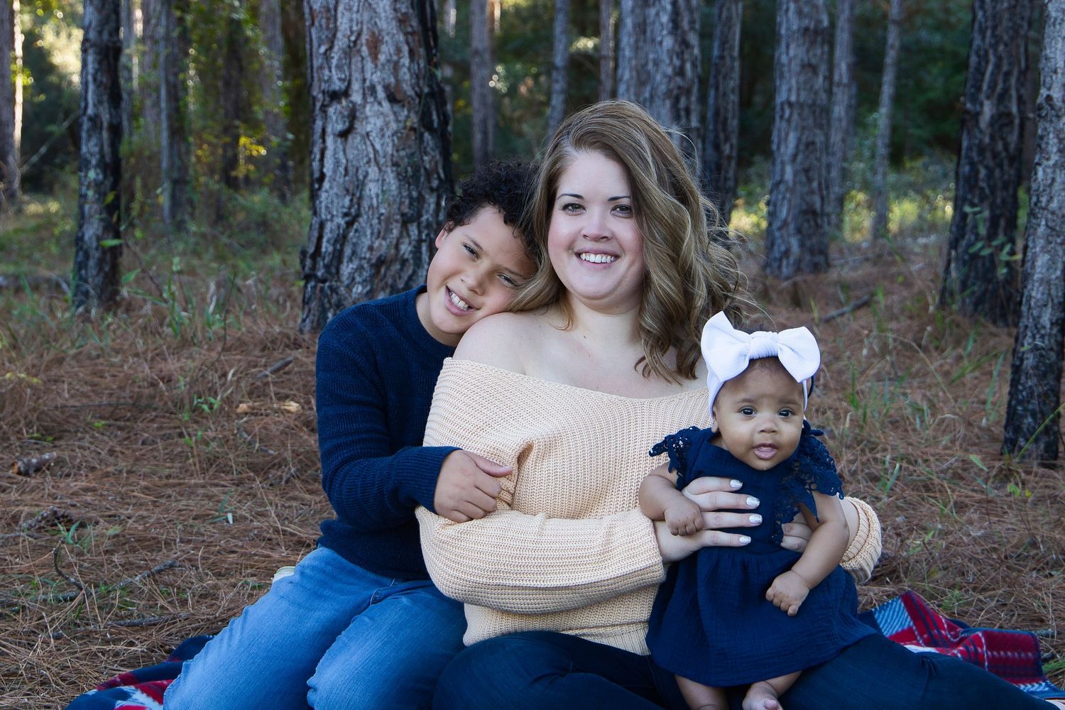 Family portrait of a smiling woman with two children sitting on a blanket in a pine forest during autumn.