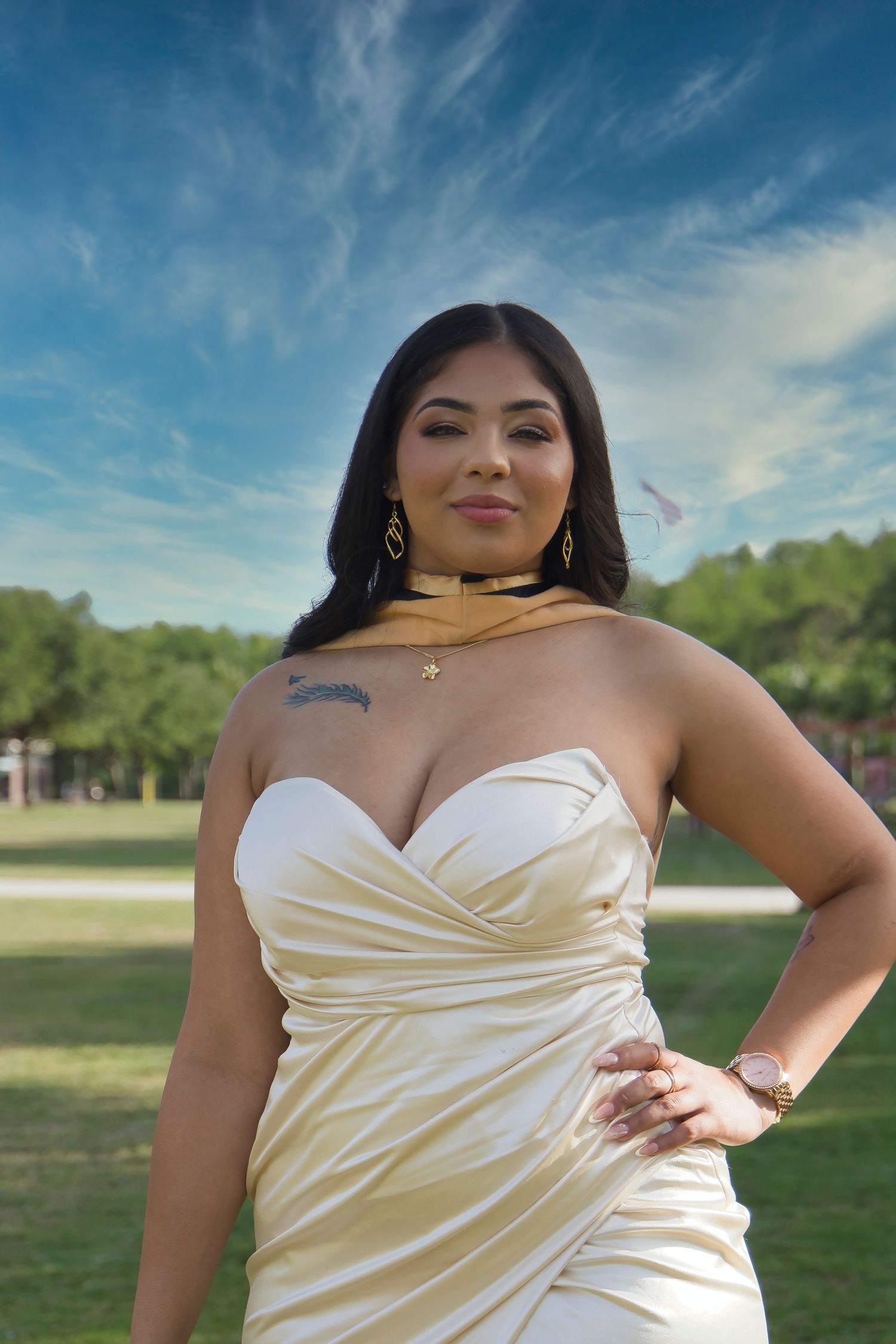 Woman in white strapless dress with gold accessories poses outdoors in a park under blue sky.
