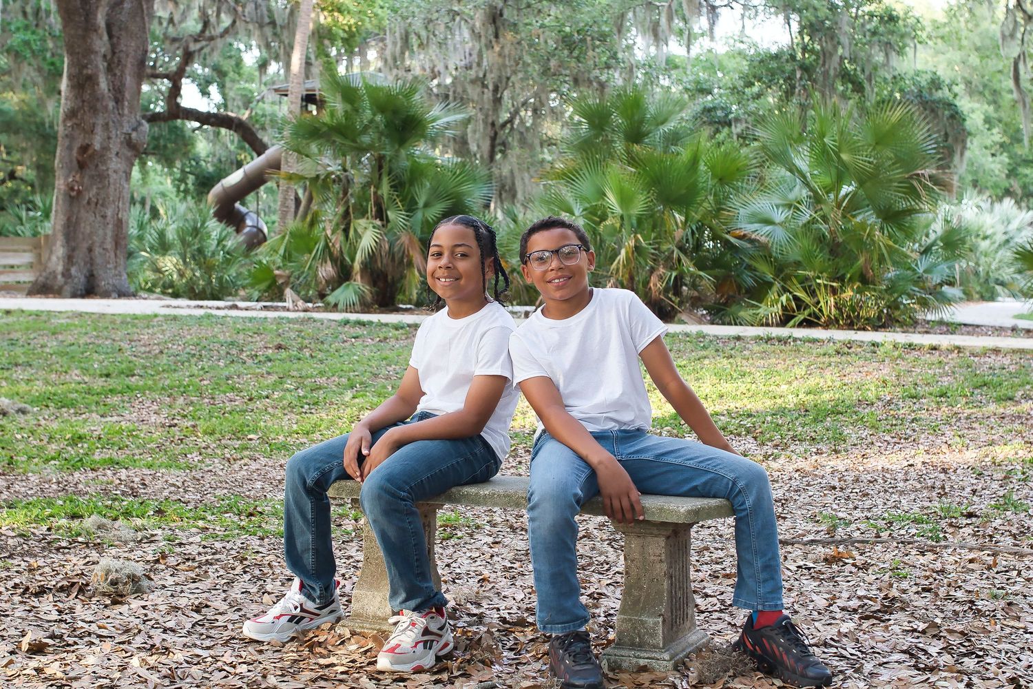 Two boys in white shirts and jeans sitting together on a stone bench in a park.