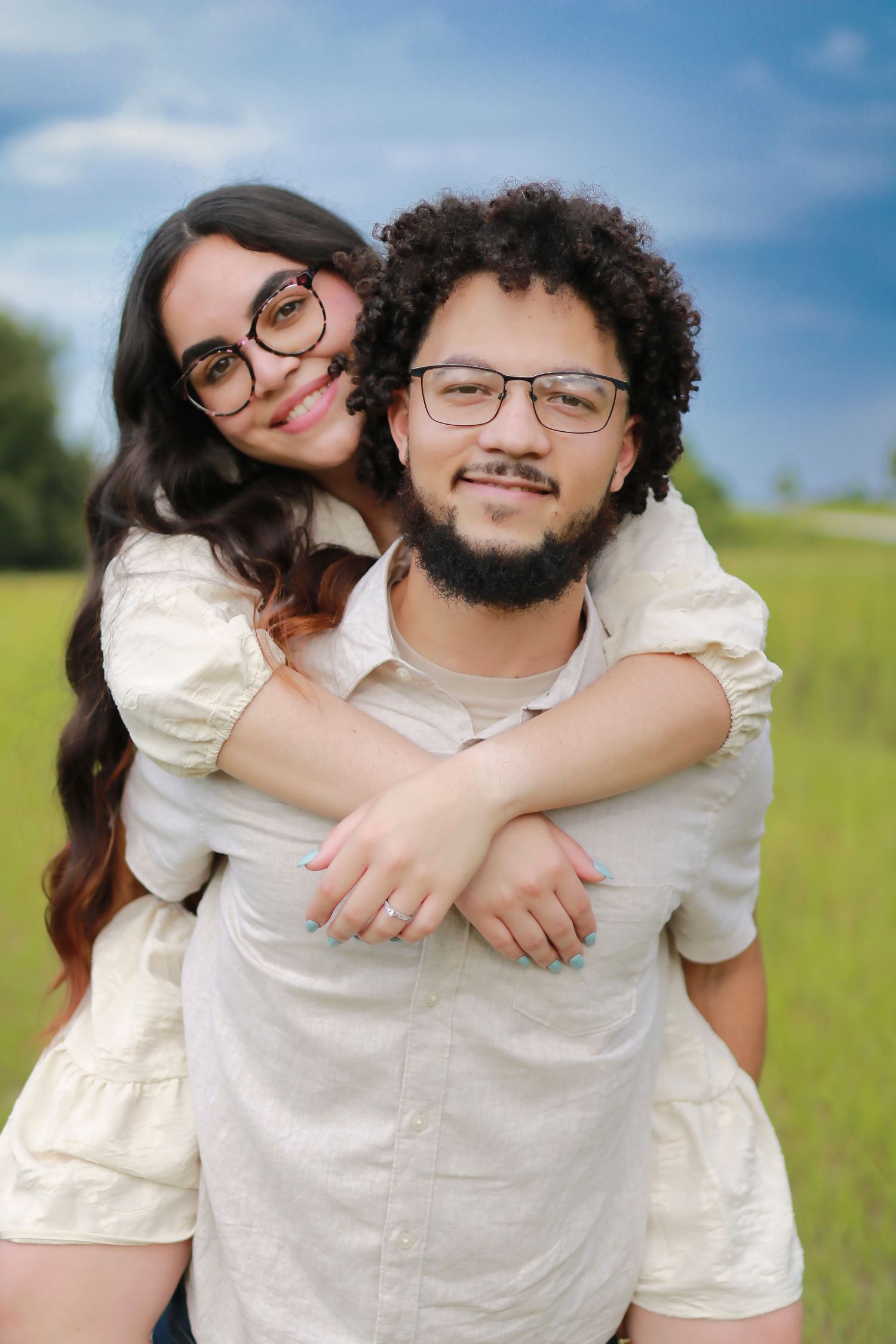 A playful couple shares candid moments together in matching white outfits against a bright blue sky and grassy field.