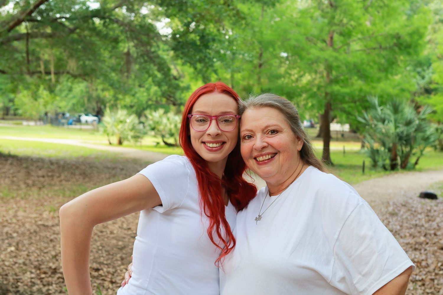 Two women smiling outdoors in white t-shirts, one with red hair and glasses, posing together in a park setting.