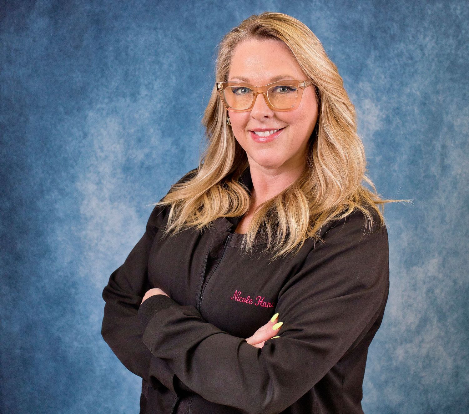 Smiling blonde woman in black embroidered scrubs with arms crossed against blue background.