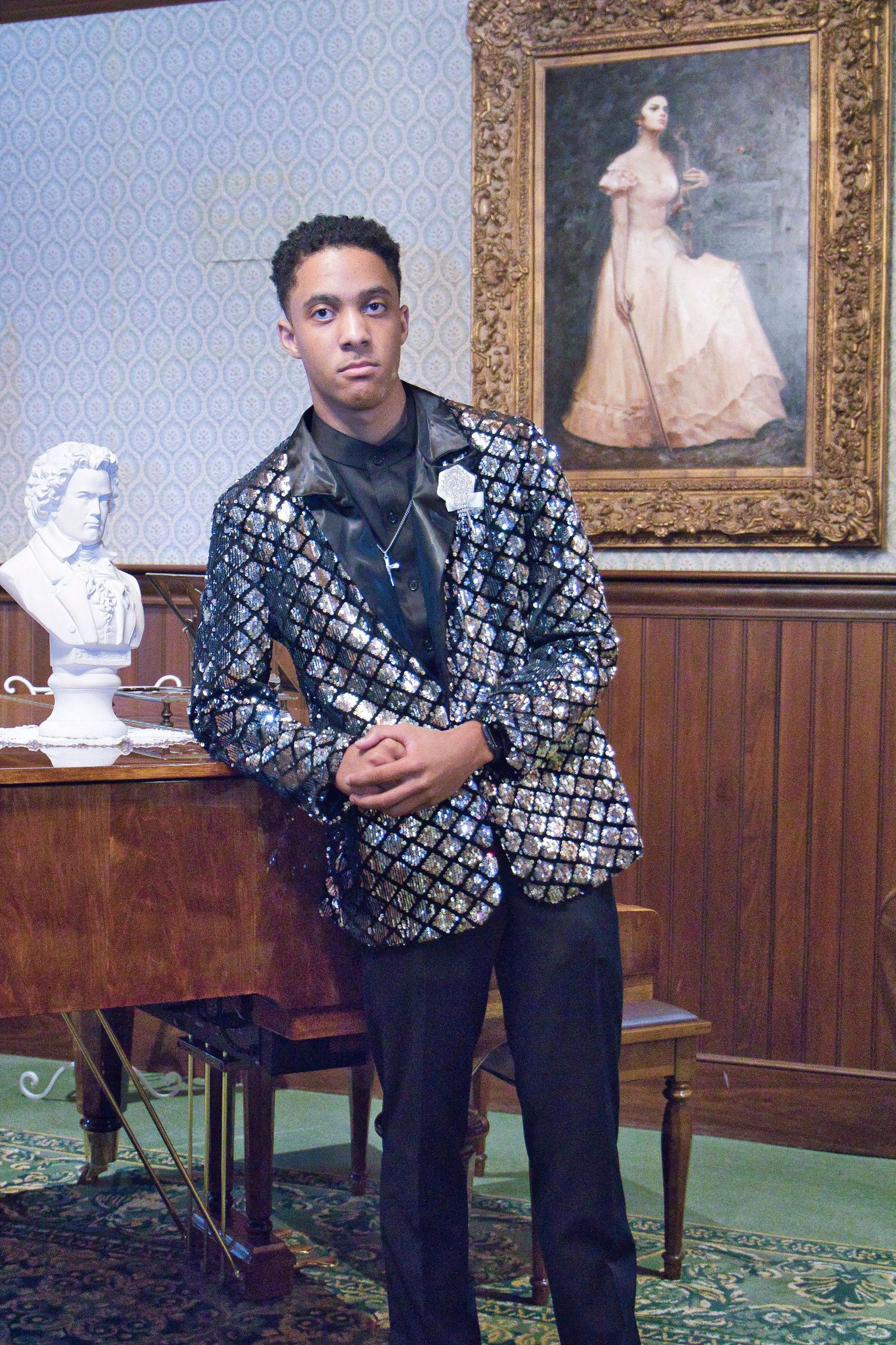 Stylishly dressed person in patterned jacket poses next to ornate framed artwork.