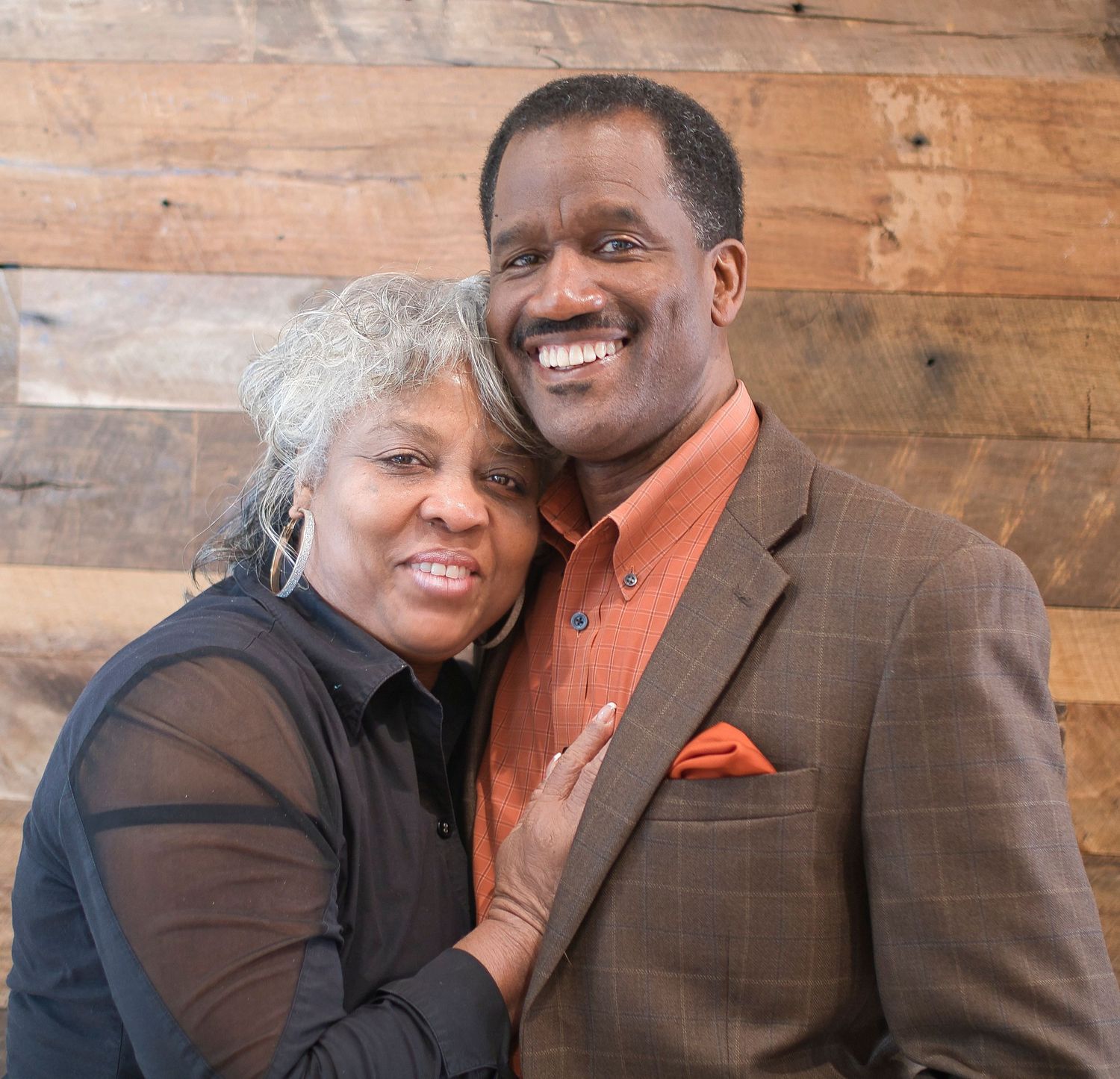 A happy couple embraces while posing against a wooden wall background.