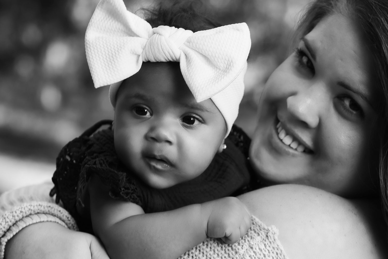 Black and white photo of a smiling woman holding a cute baby girl wearing a large white bow headband.