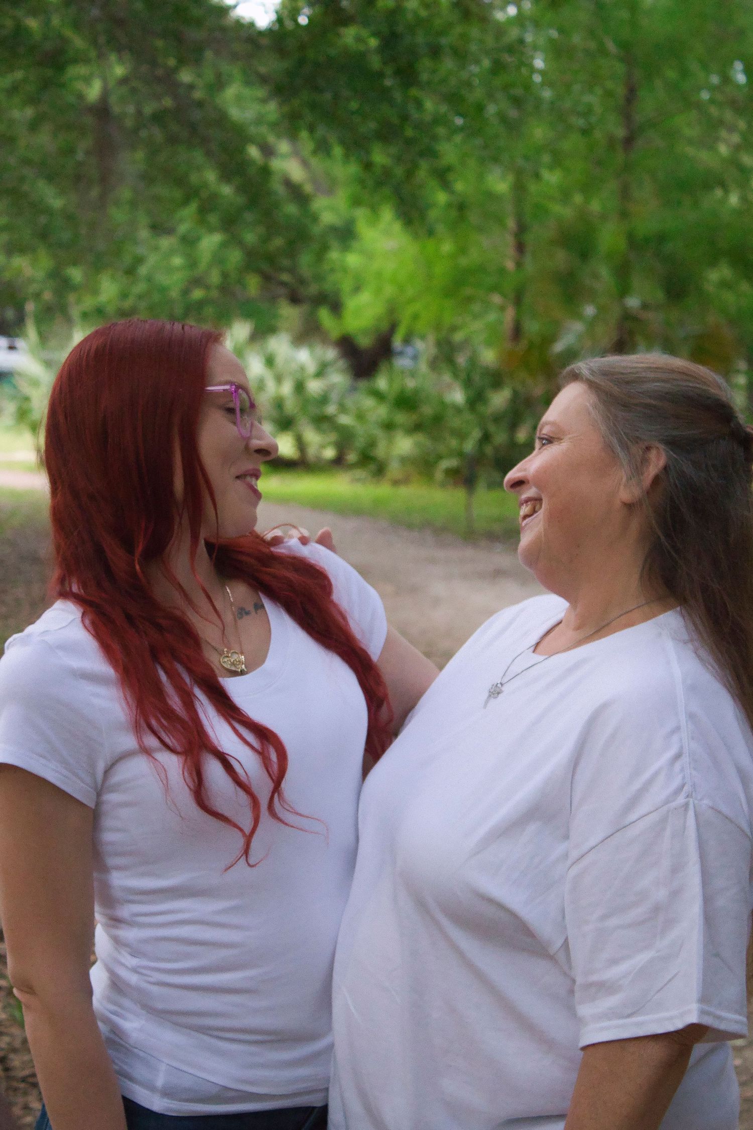 Two women in white t-shirts smile at each other outdoors, one with red hair and glasses, surrounded by lush green trees.