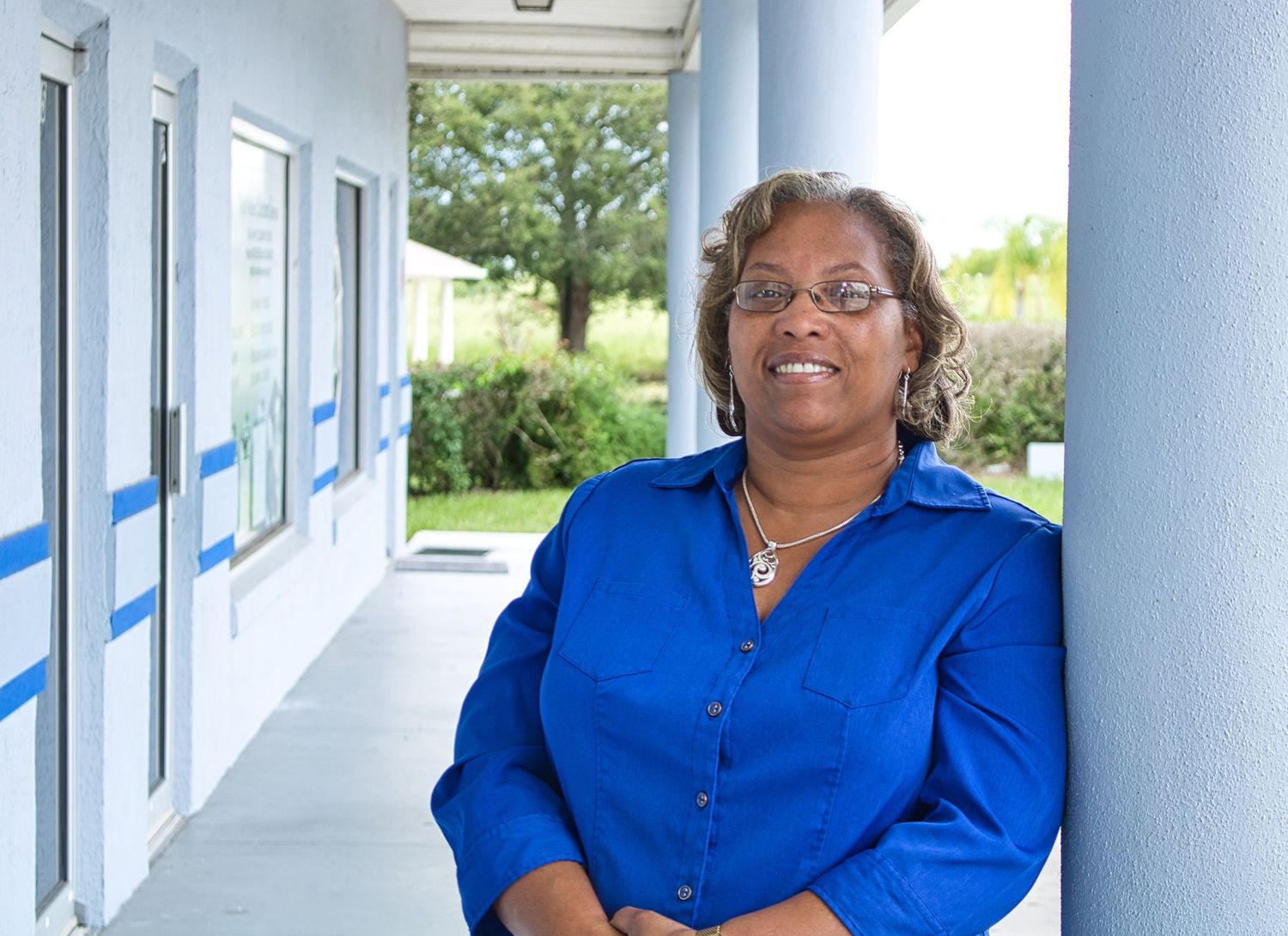A smiling woman in a blue shirt leans against a column in a bright outdoor hallway with greenery in the background.