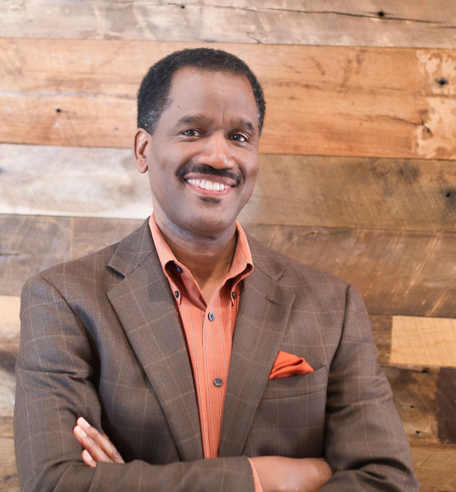 Smiling man in brown blazer and orange shirt with pocket square, arms crossed, posed against rustic wood wall.