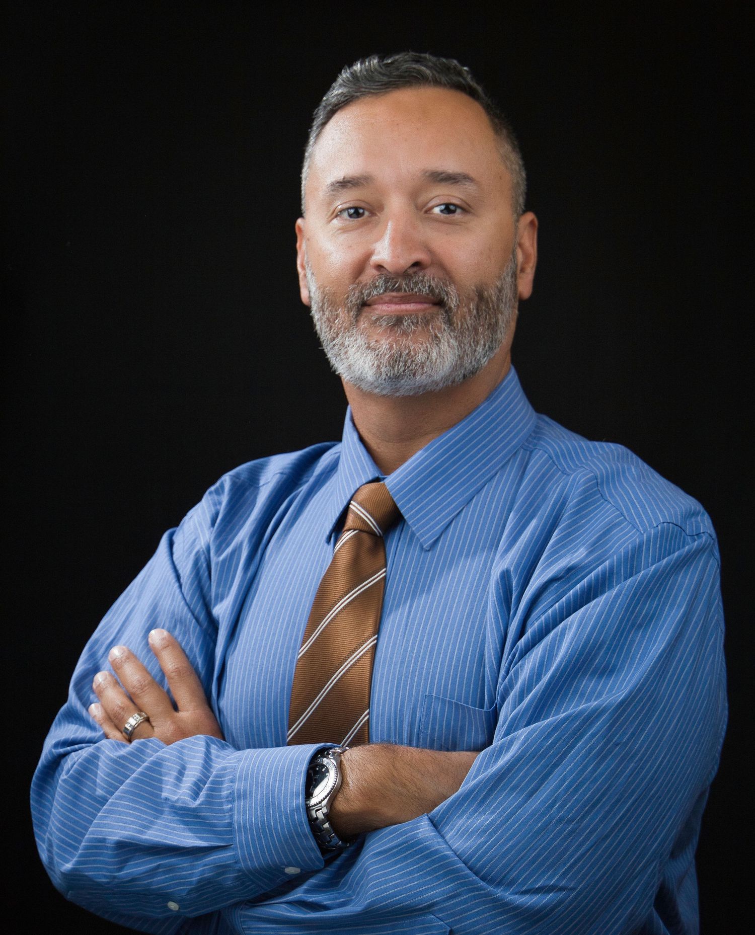 Professional headshot of person in blue shirt and brown tie against dark background.