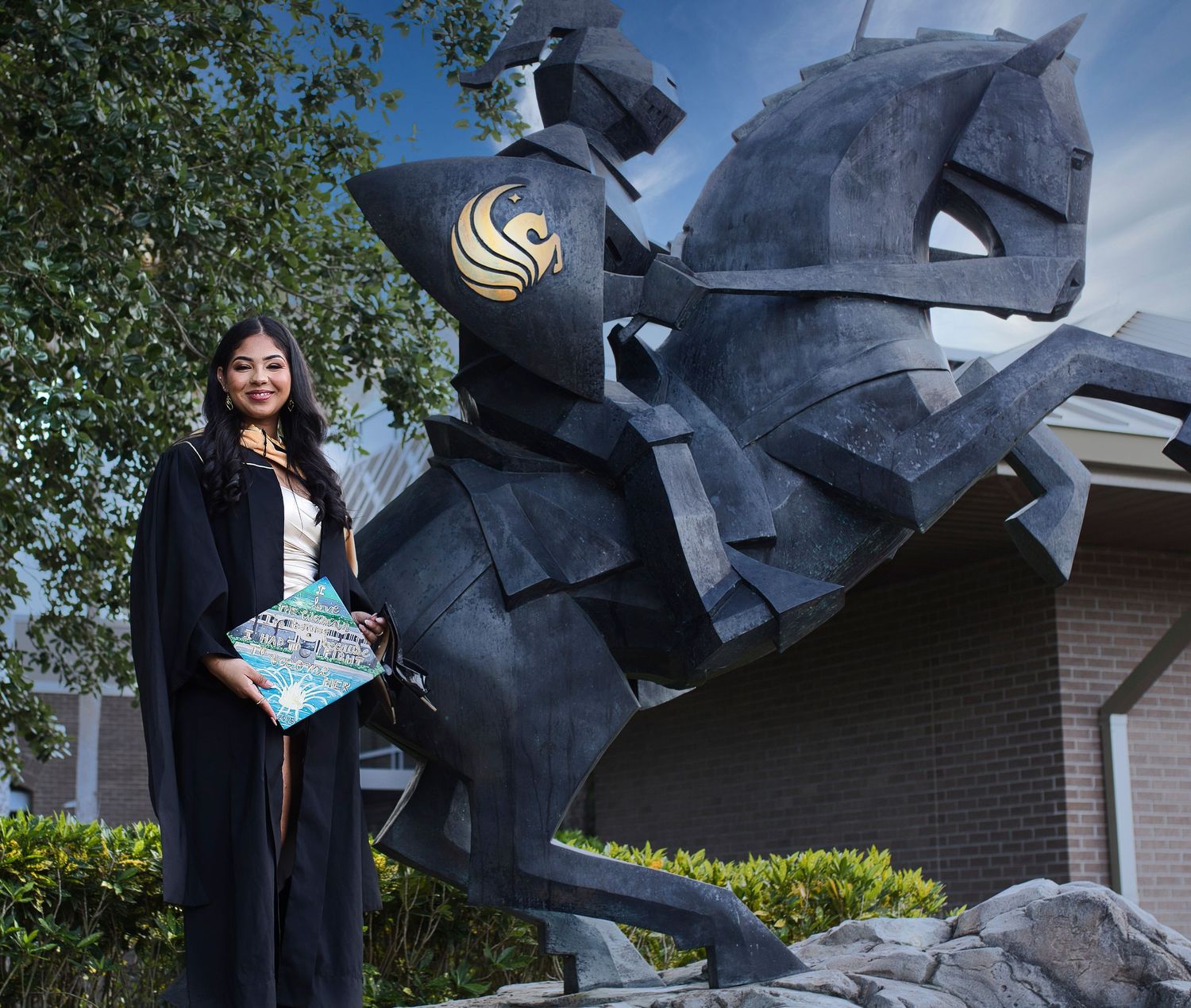 A graduate in academic regalia stands beside a large dark metal knight sculpture on horseback at a university campus.
