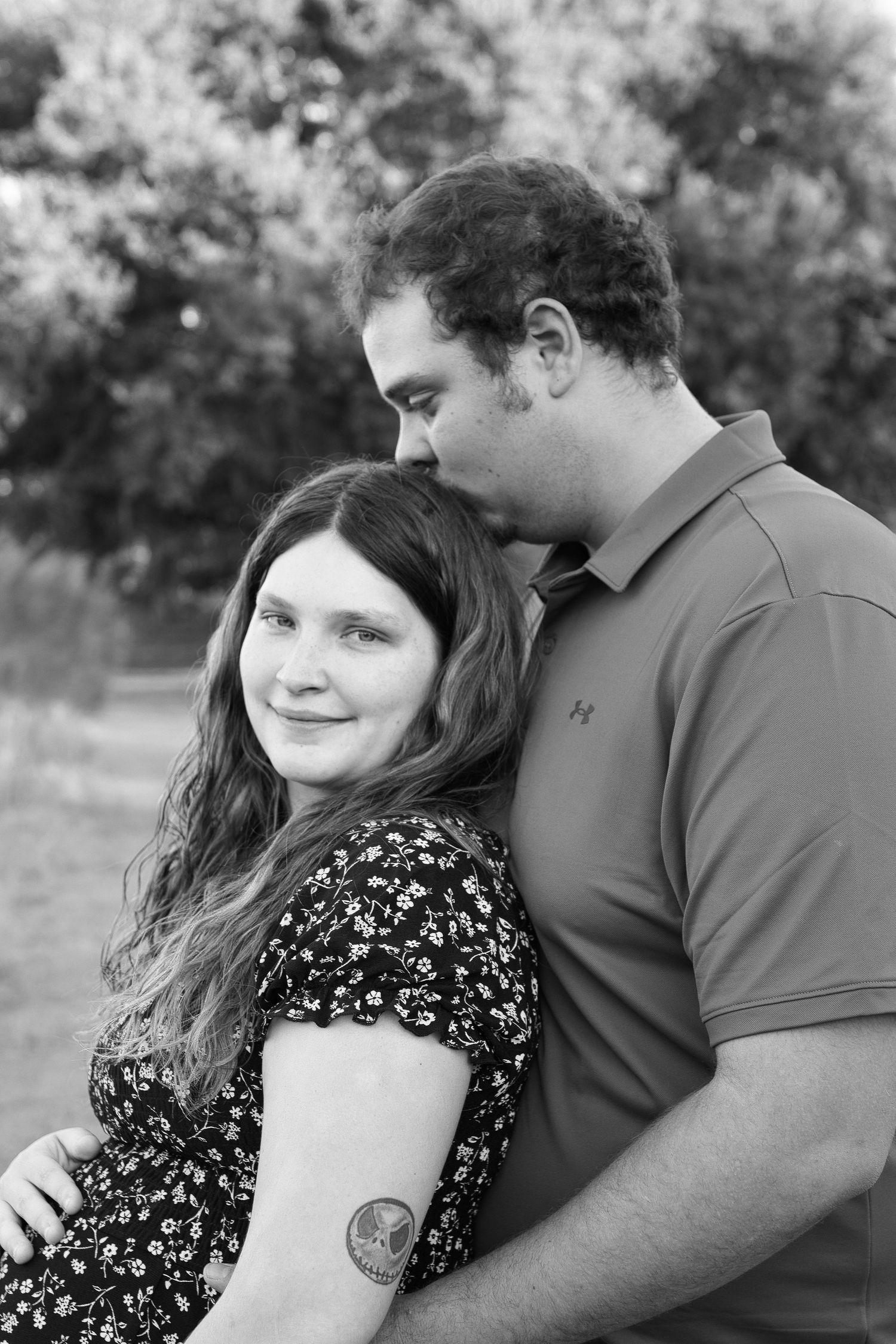 Black and white photo of a couple embracing outdoors, man kissing woman's head, woman smiling with floral dress and arm tattoo.