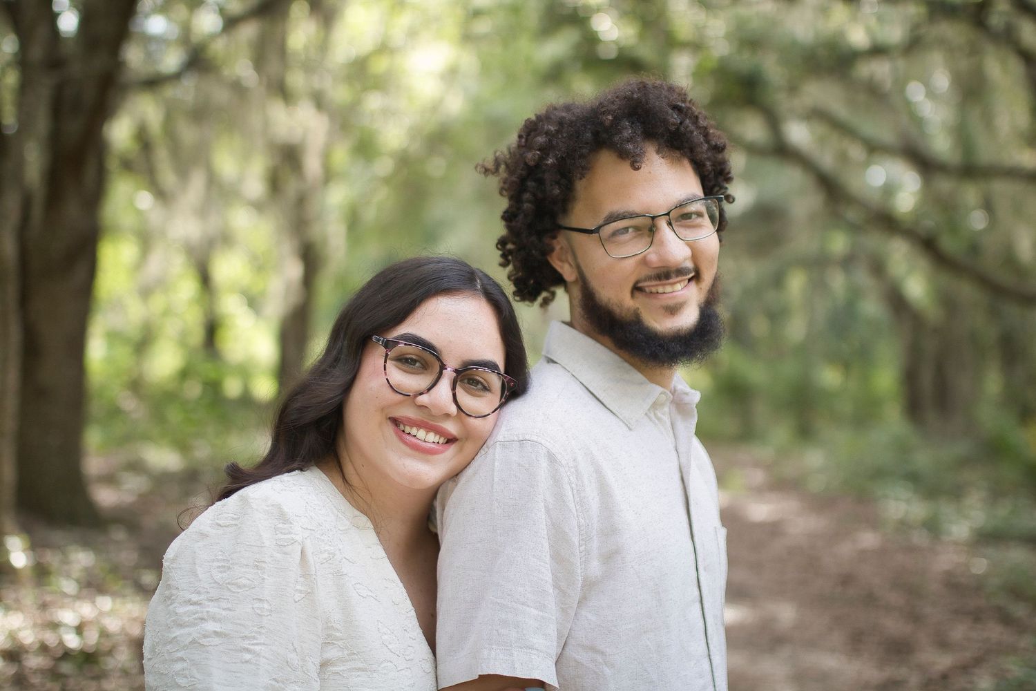 A smiling couple wearing glasses poses outdoors among moss-covered trees in a lush green forest setting.