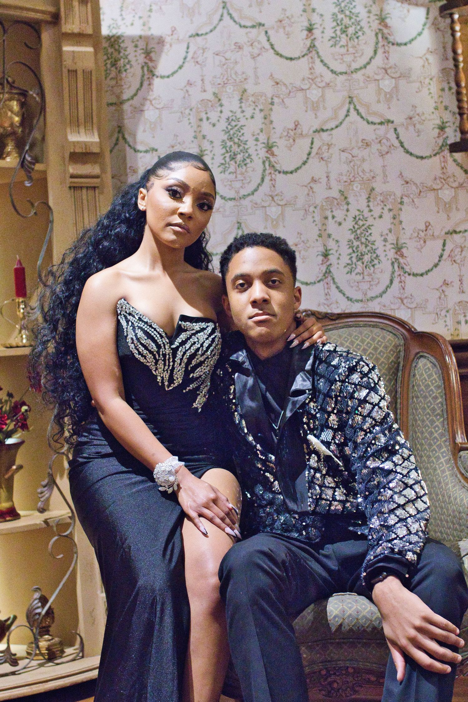 Elegant couple posing in formal attire, woman in black beaded gown and man in embroidered blazer, ornate vintage interior.