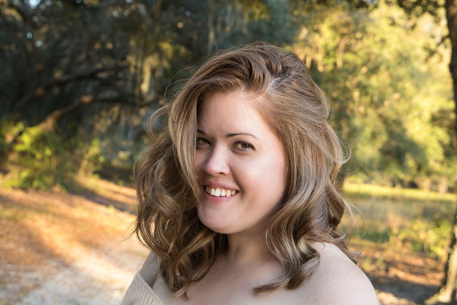 Smiling young woman with wavy blonde hair posing outdoors on a sunlit dirt path surrounded by trees.
